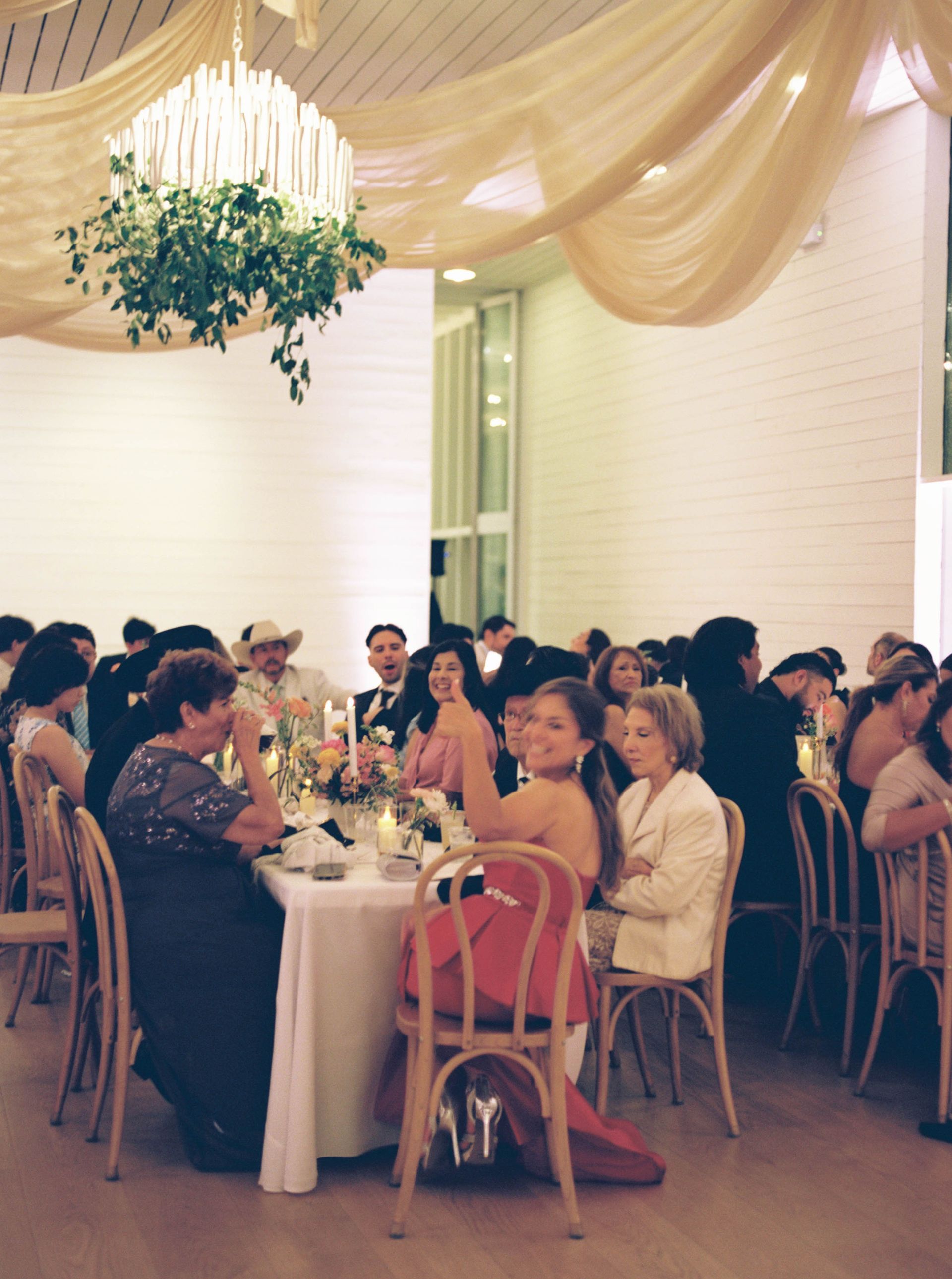 Wedding reception: Guests seated at tables in a bright room, some waving or looking at camera.