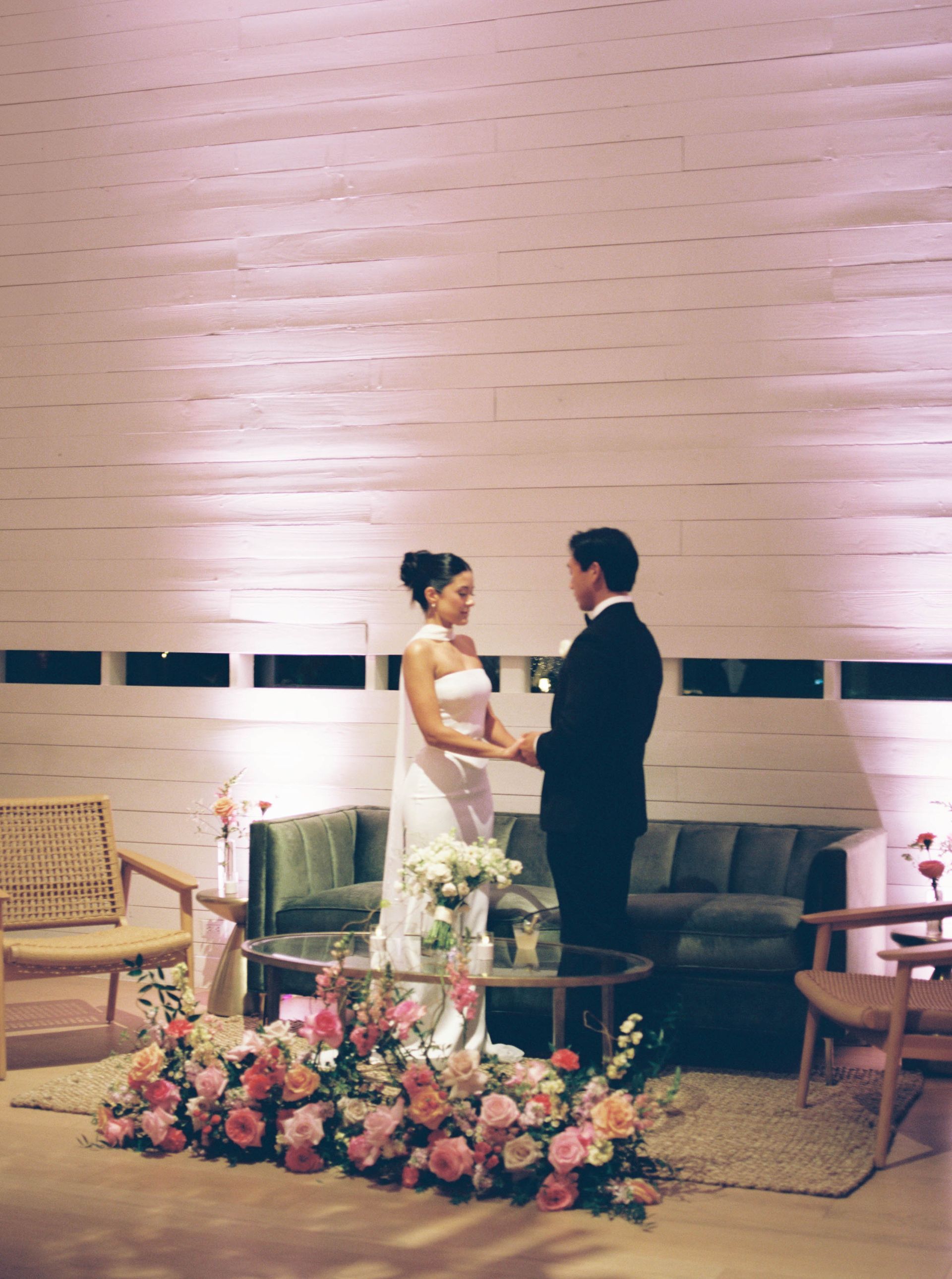 Bride and groom holding hands, exchanging vows in a modern setting with floral decor and a green couch.