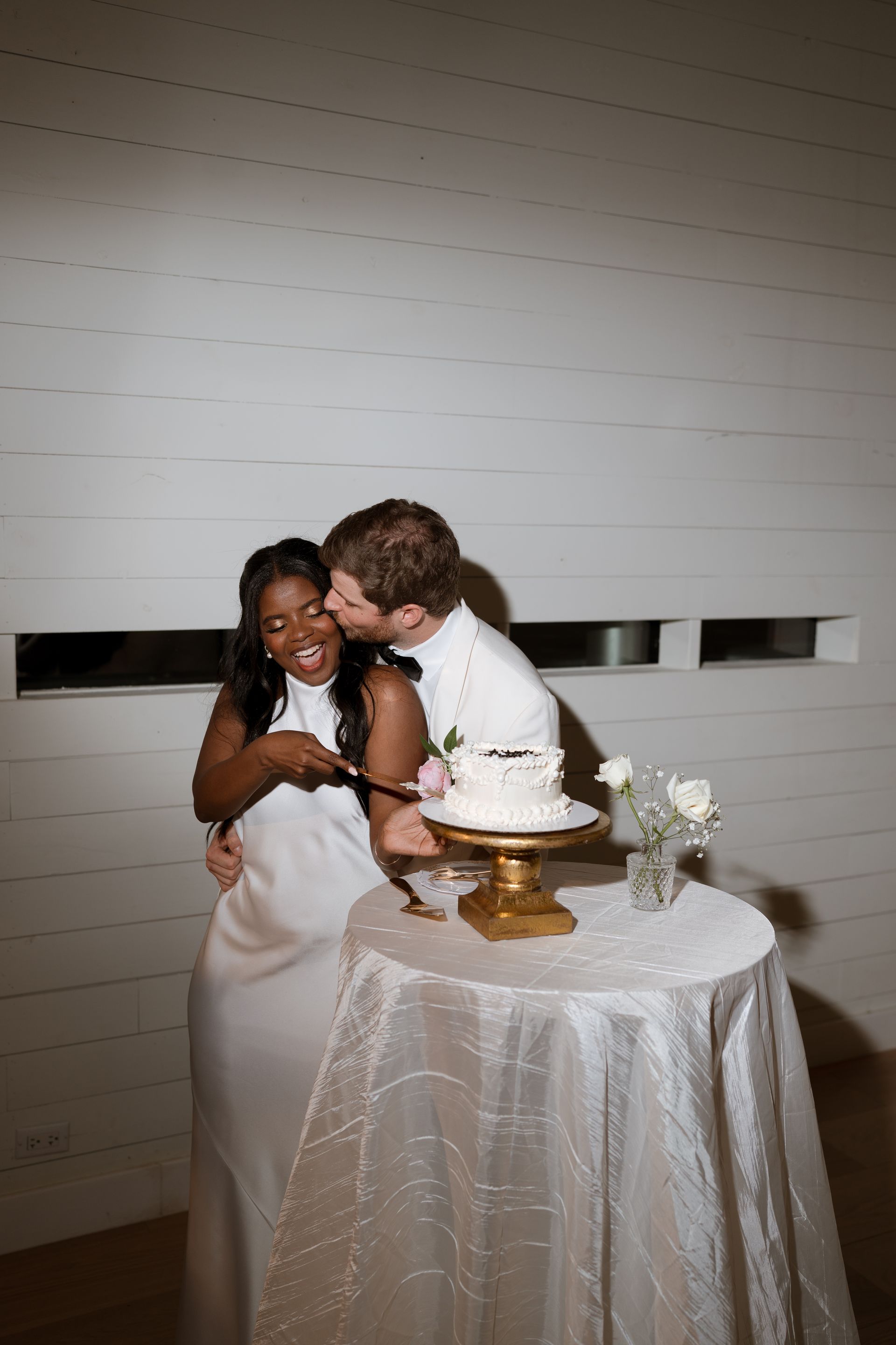 Newlyweds cutting cake together. Bride laughs, groom kisses her cheek. White walls and table, gold cake stand.
