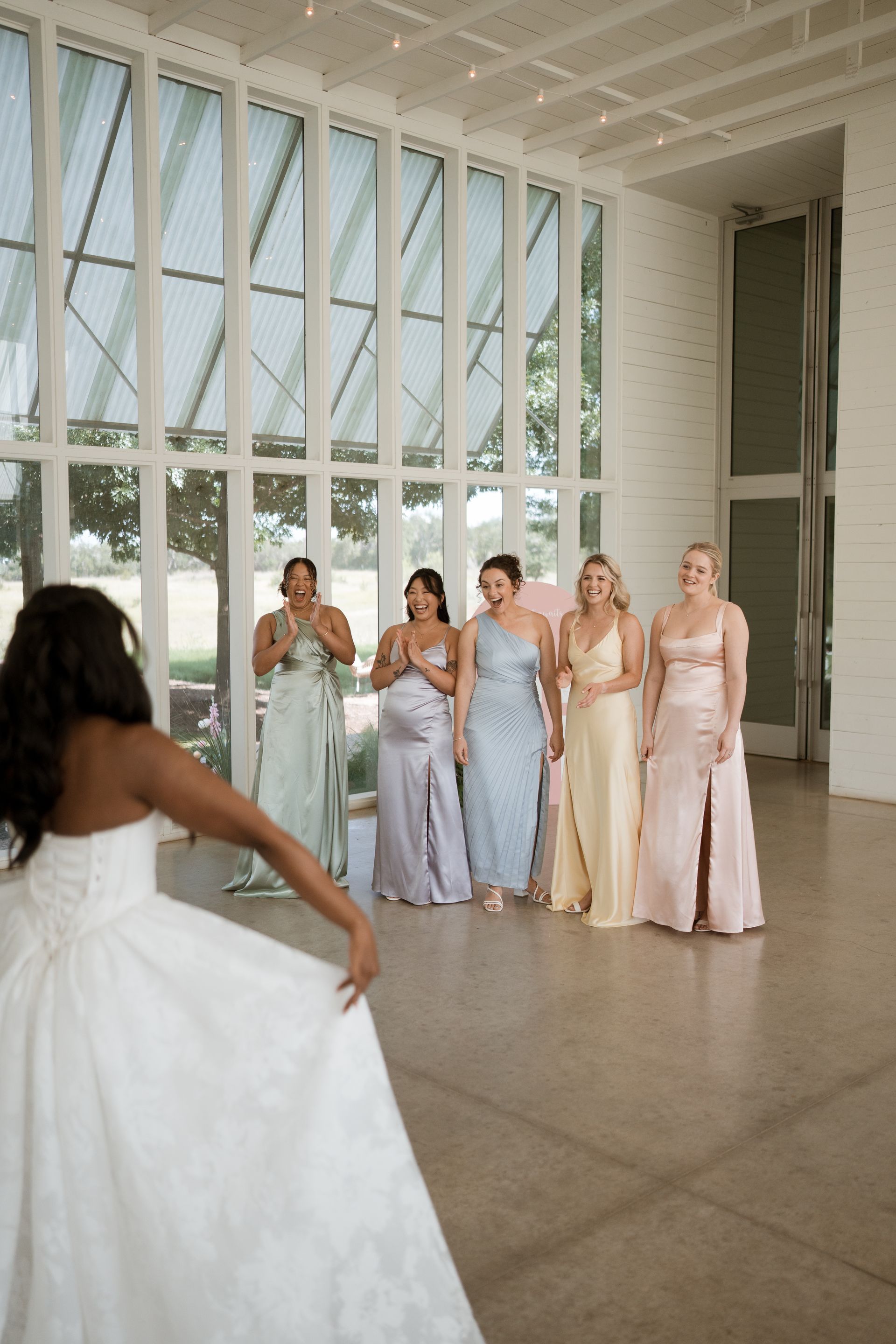 Bride in white dress twirls, facing six bridesmaids in pastel gowns, inside bright room with large windows.