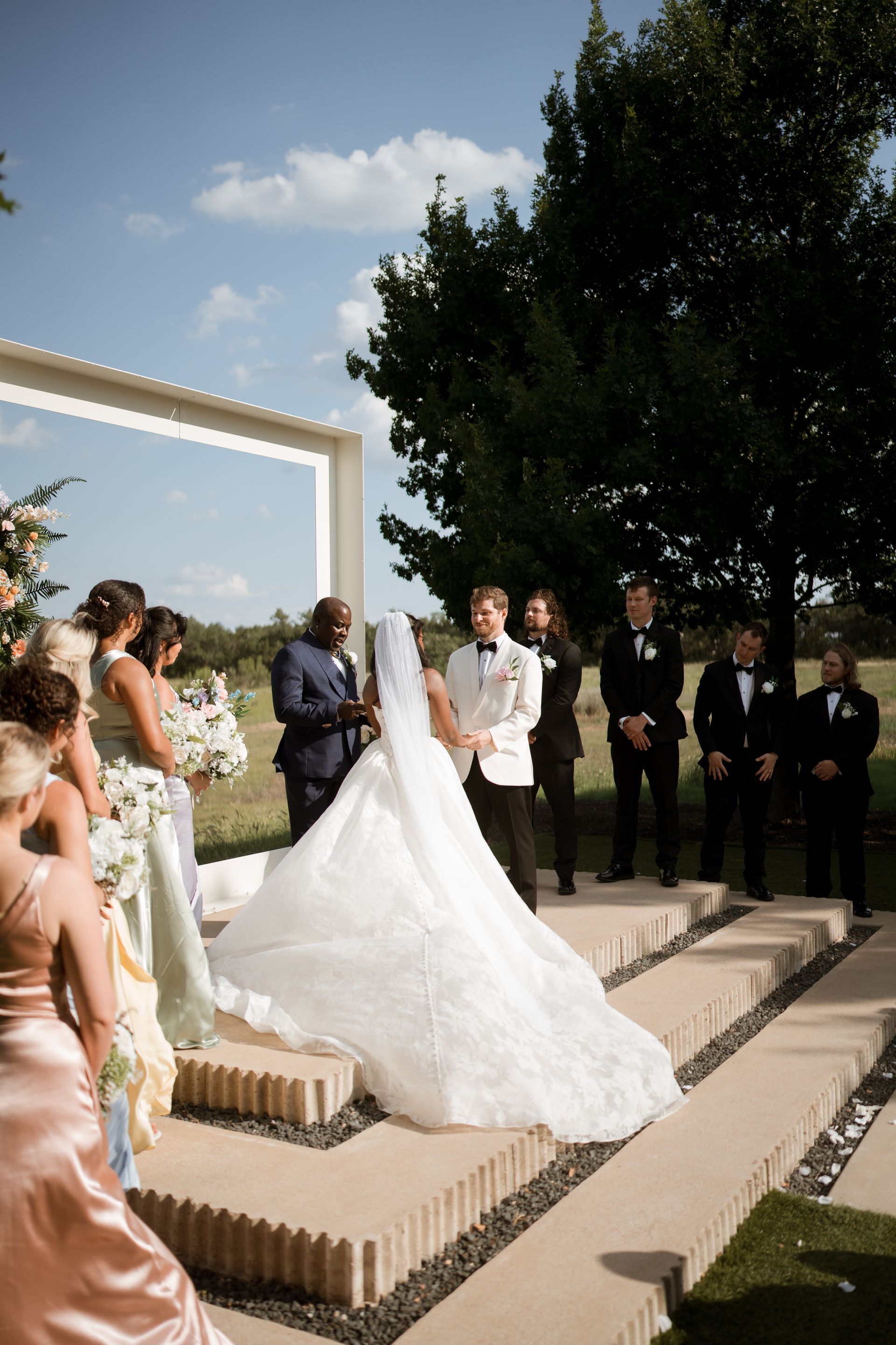 Wedding ceremony: Bride in white gown, groom, officiant, and wedding party outdoors.