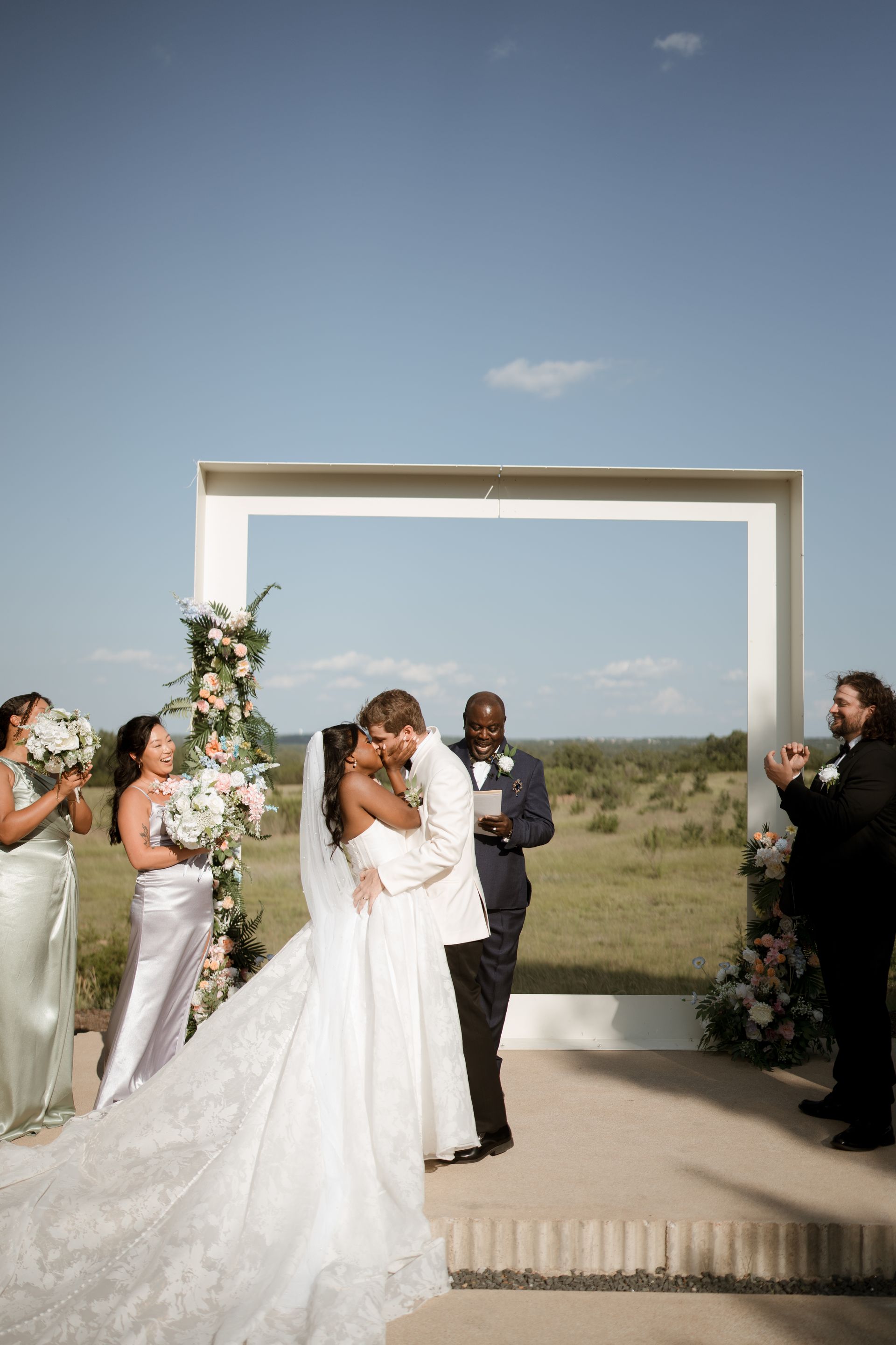 A bride in white dress walks with her father down a path, leading to a building with glass walls.