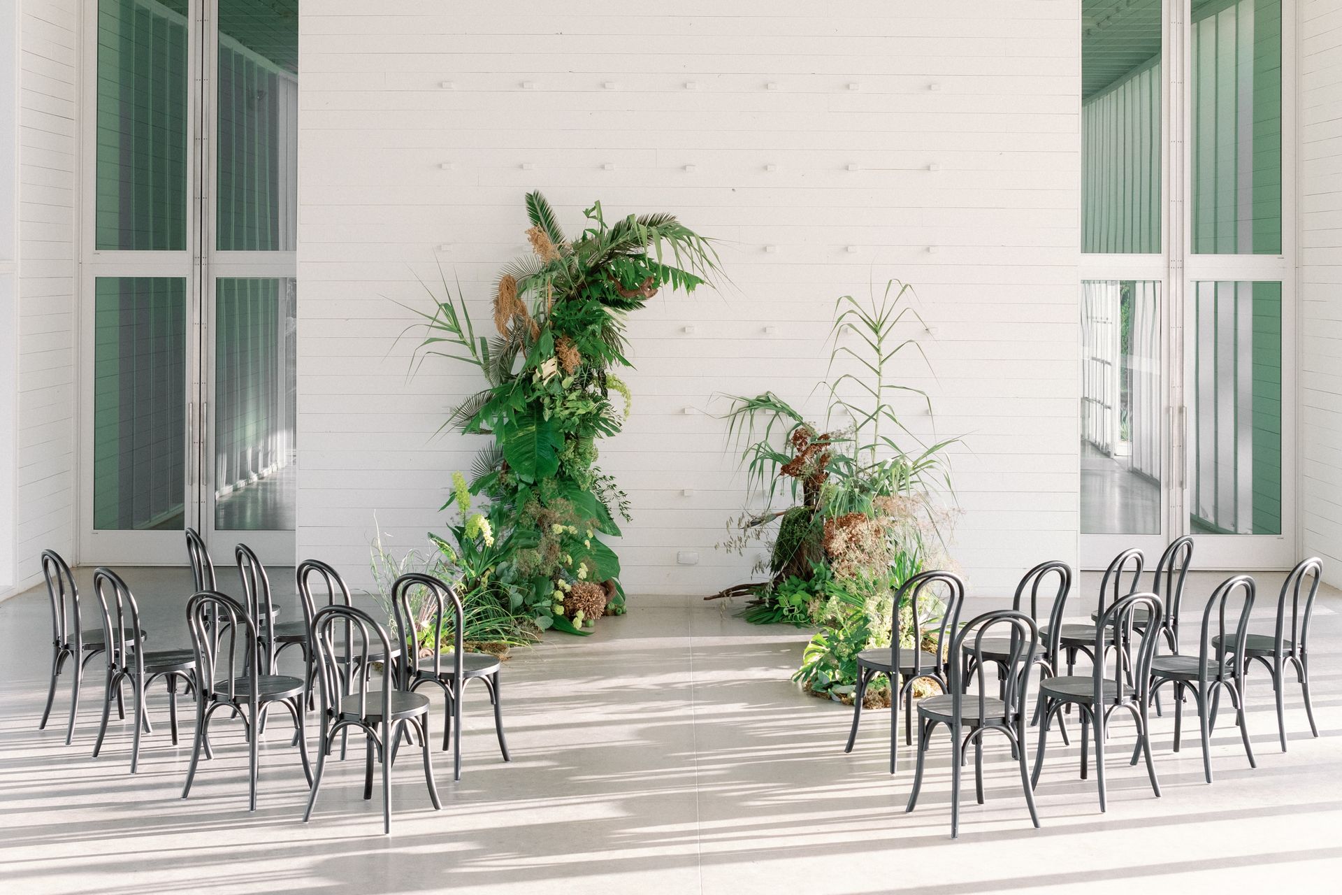 Wedding ceremony setup. Black chairs face a floral arch against a white wall.