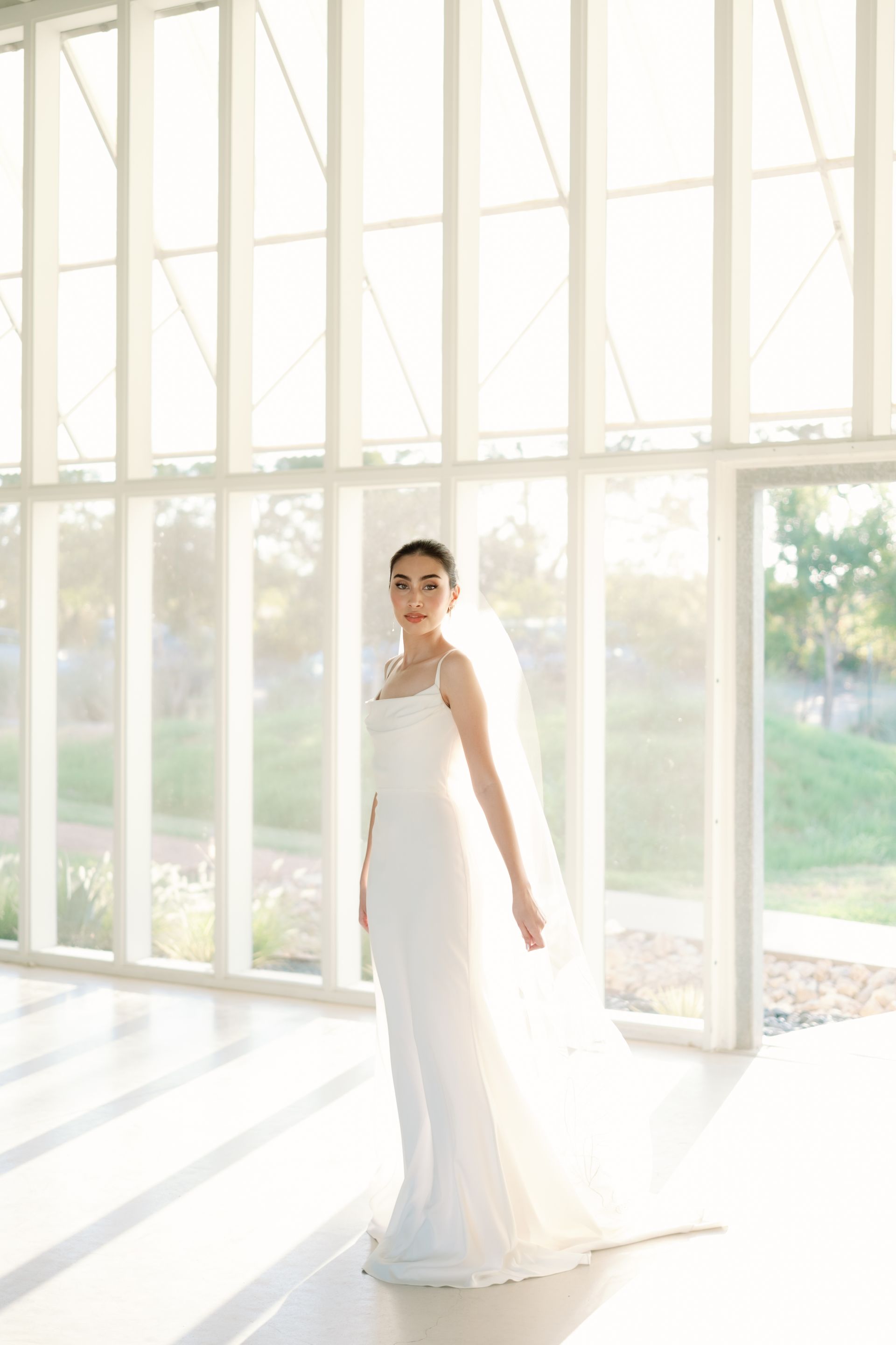 Bride in a white gown stands in a bright, modern space, veil trailing.