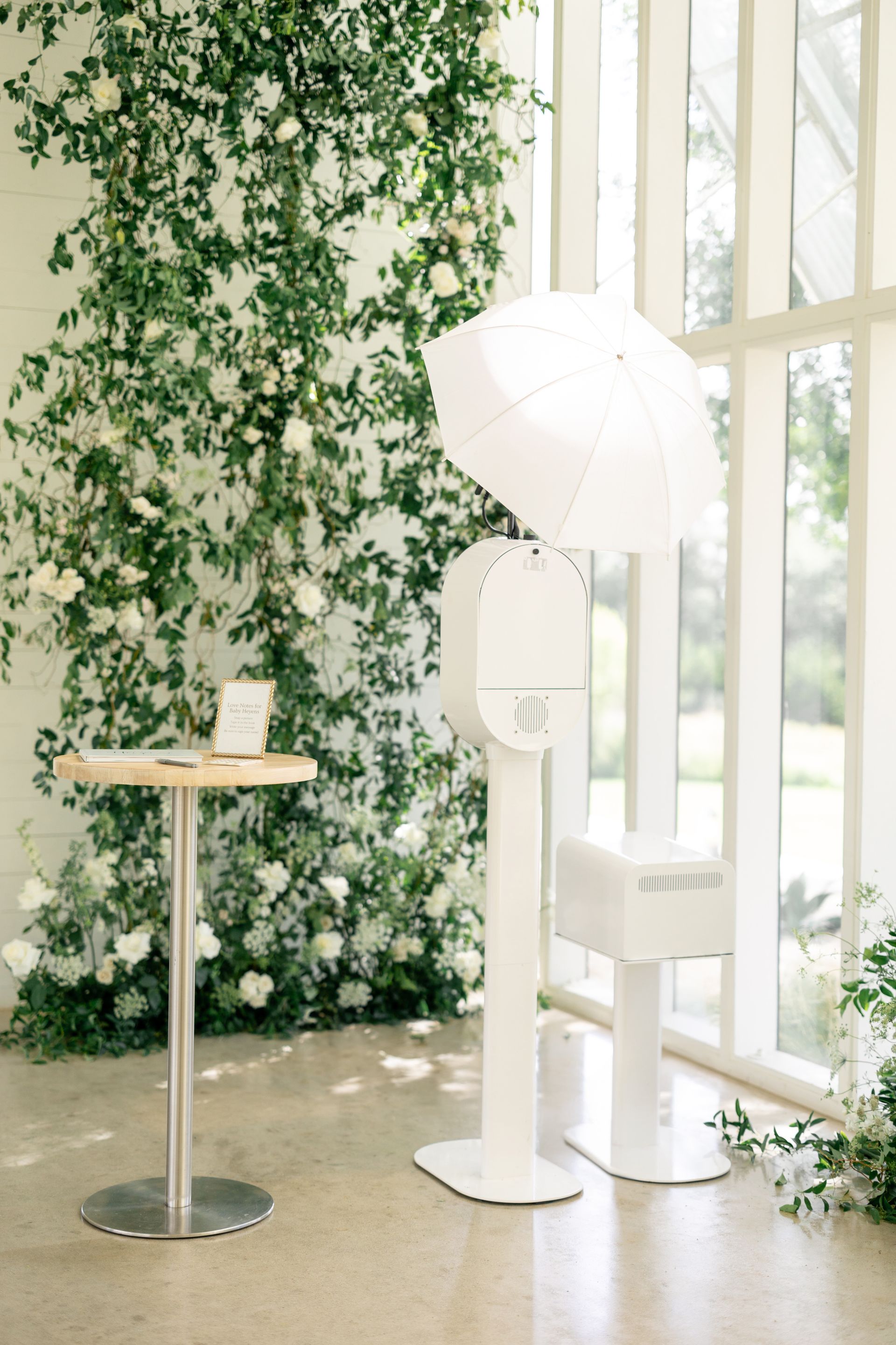 White photo booth with umbrella near a floral wall and a small table; bright, indoor setting.