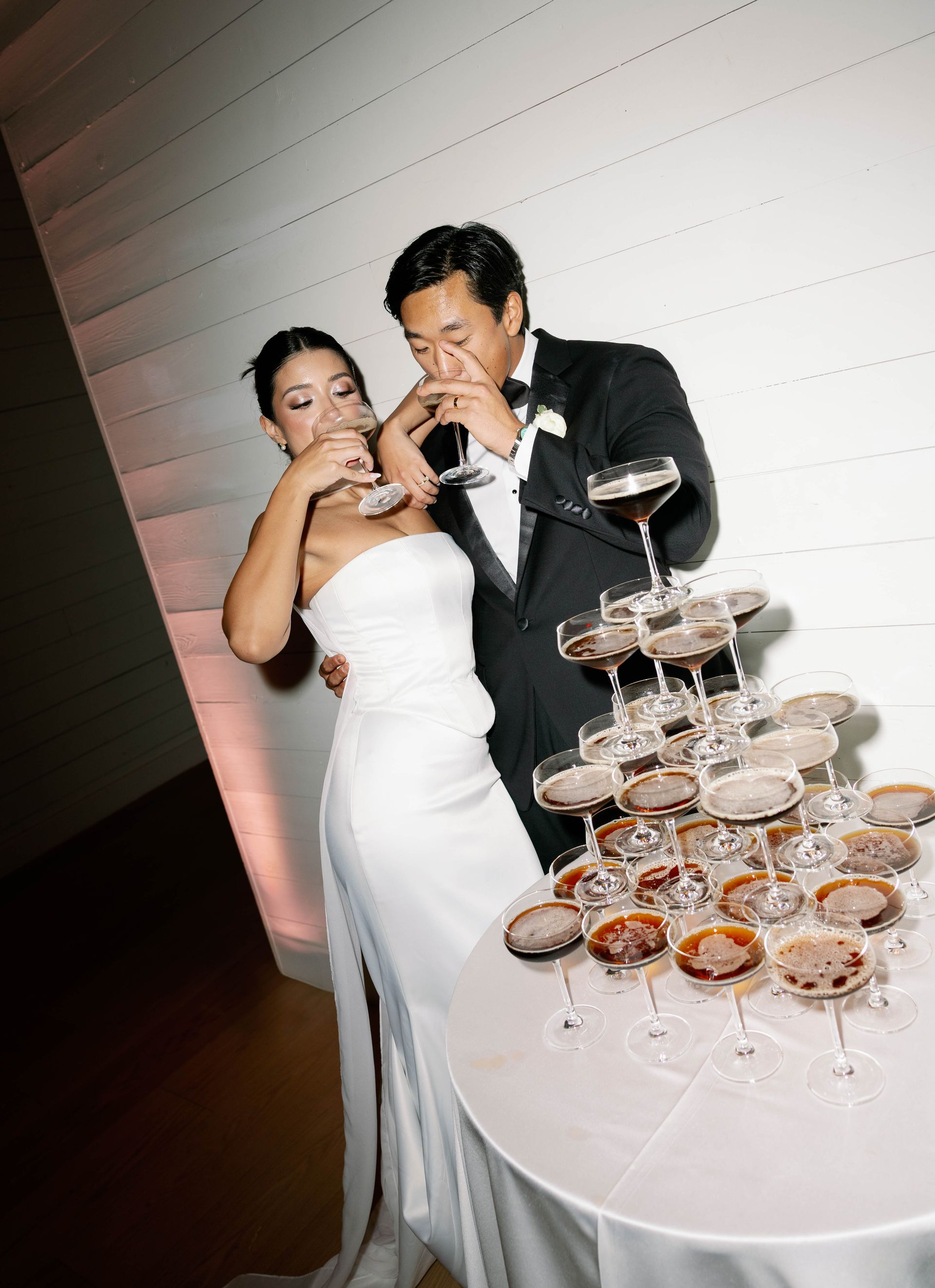 Bride and groom toast with champagne near a multi-tiered glass tower. Formal attire, white wall, celebration.