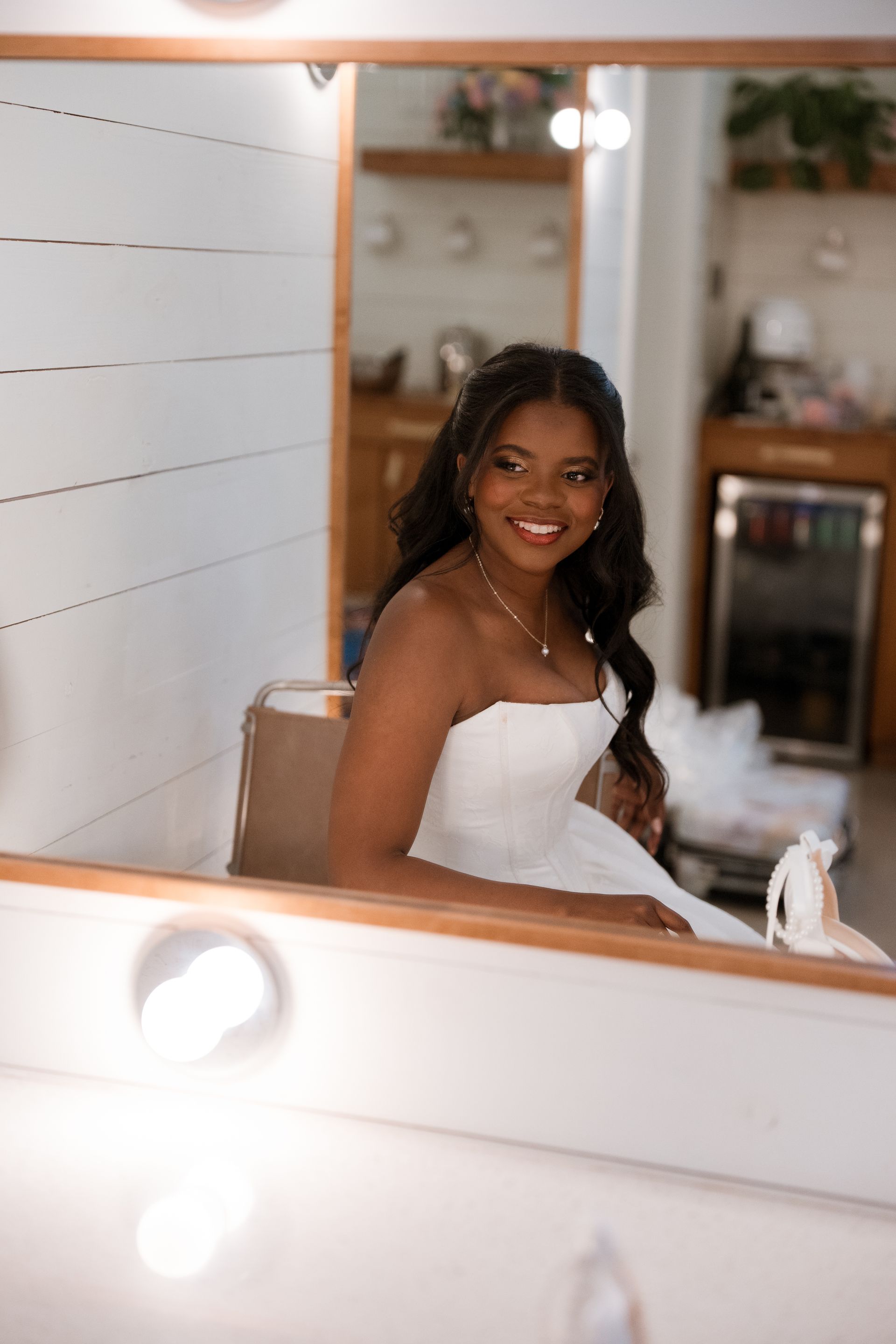 Bride with dark hair in a white strapless dress smiles, reflected in a mirror.