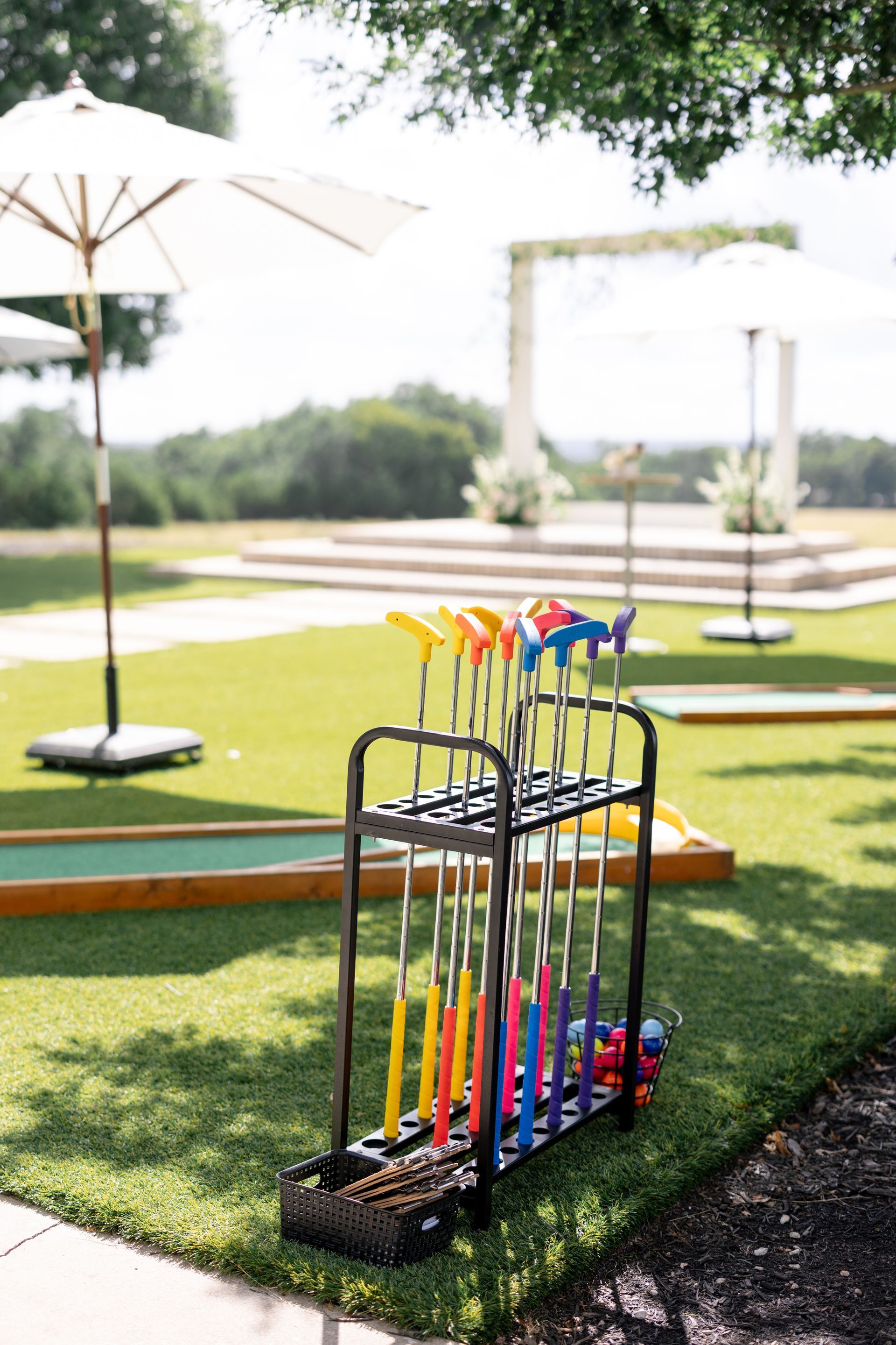 Golf putters in a black rack on green grass, with an open air wedding setting in background.