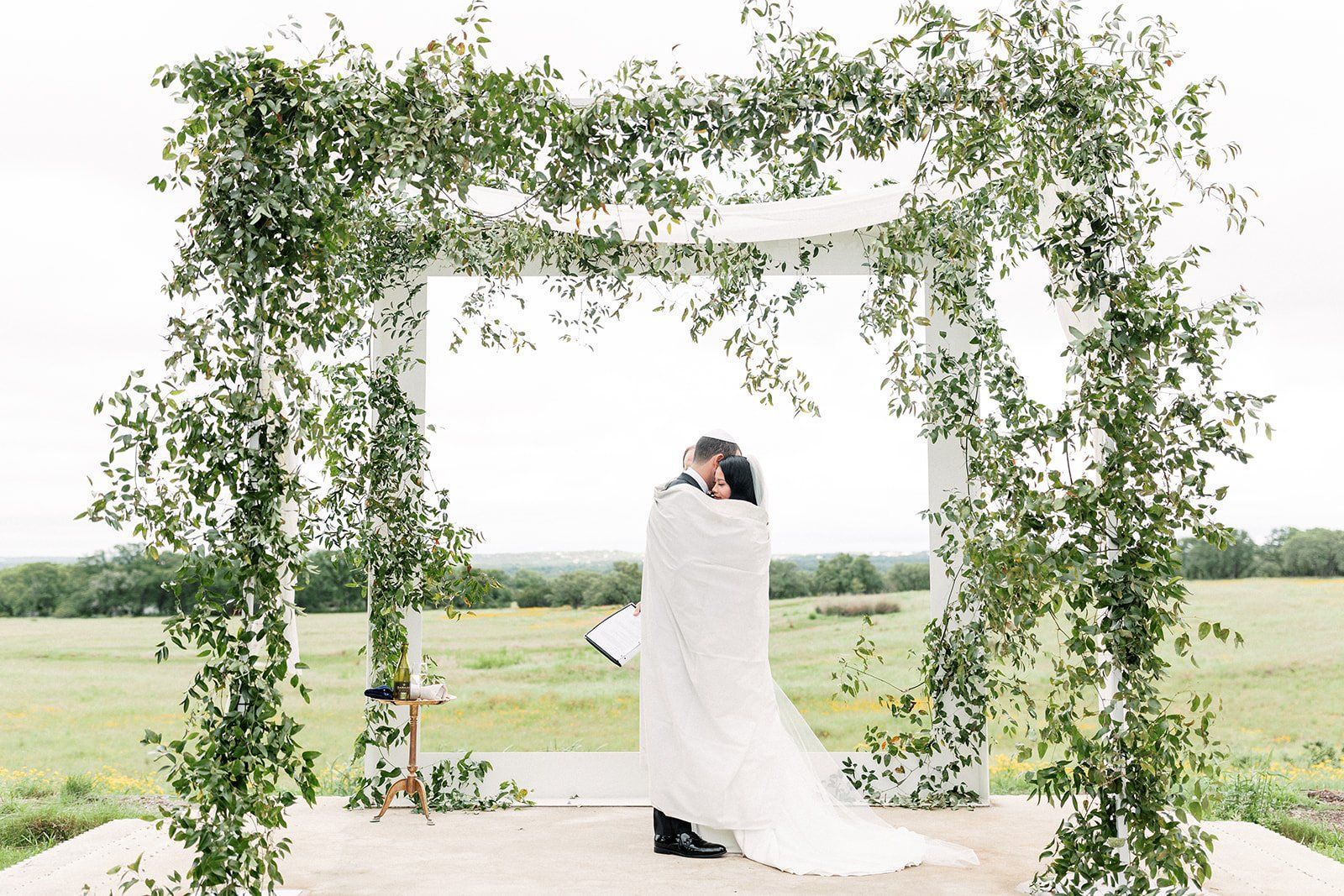 Couple embracing under a white chuppah adorned with greenery, set in a field.