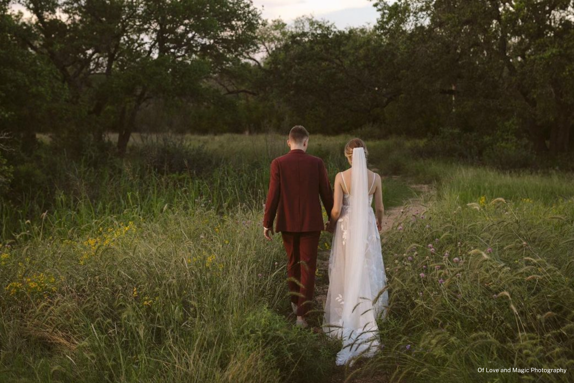 A bride and groom (who is wearing a maroon colored suit) are walking through a field holding hands with their backs to the camera. 