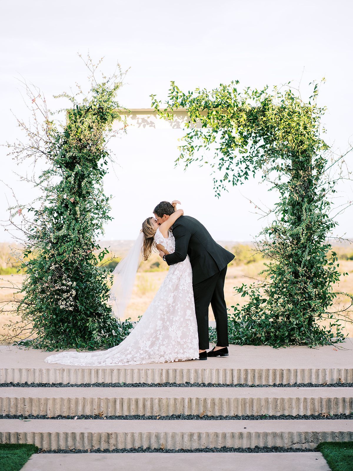 A bride and groom are kissing under a green ceremony arch.