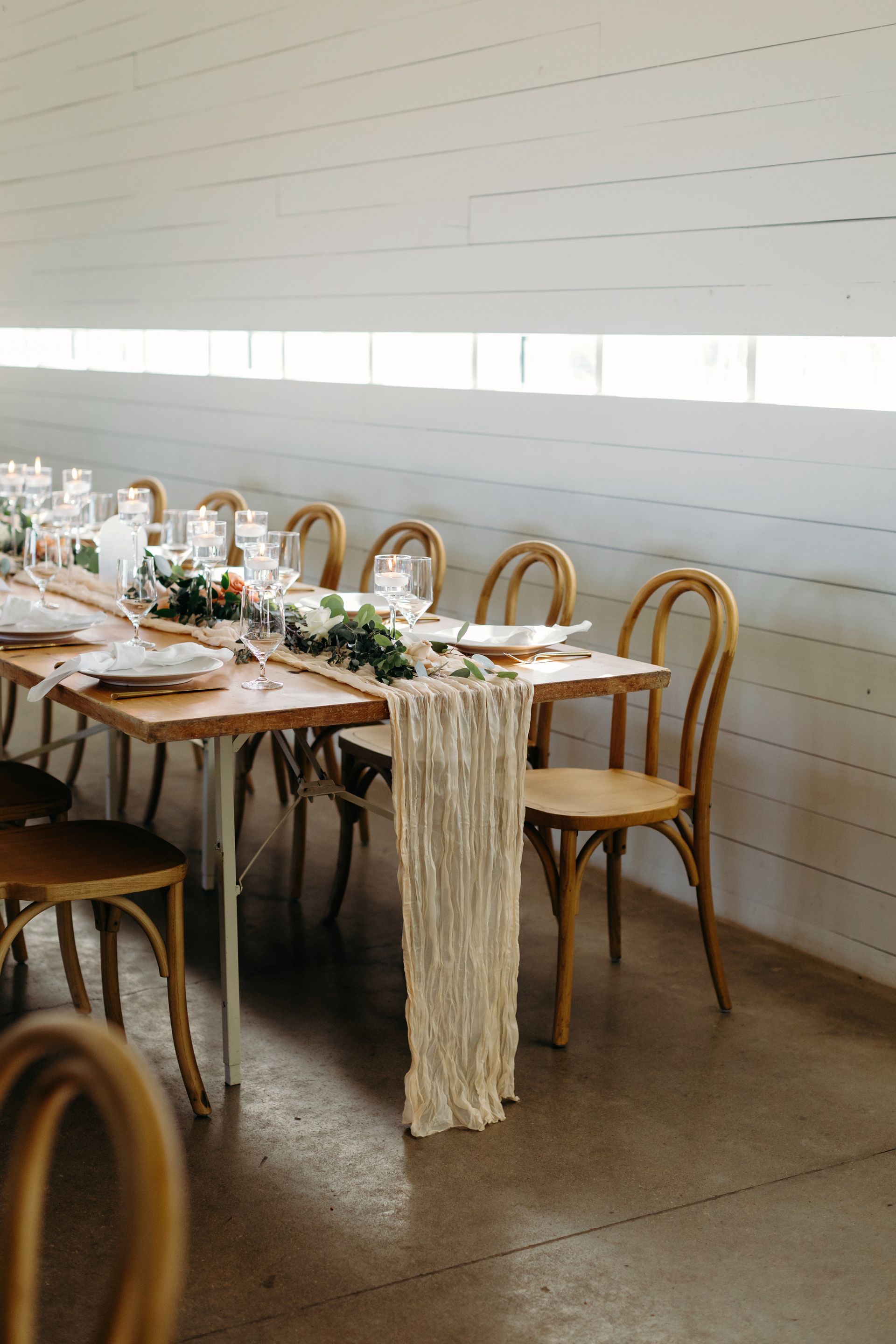 Long wooden dining table set for a celebration. Chairs, greenery, and a textured runner.