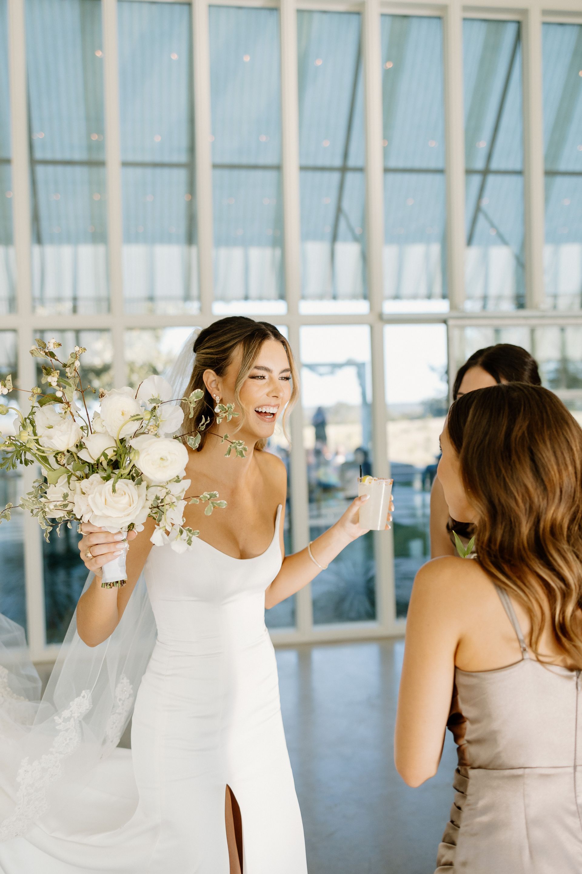Bride in white dress laughs, holding bouquet; two bridesmaids near her. Modern building.