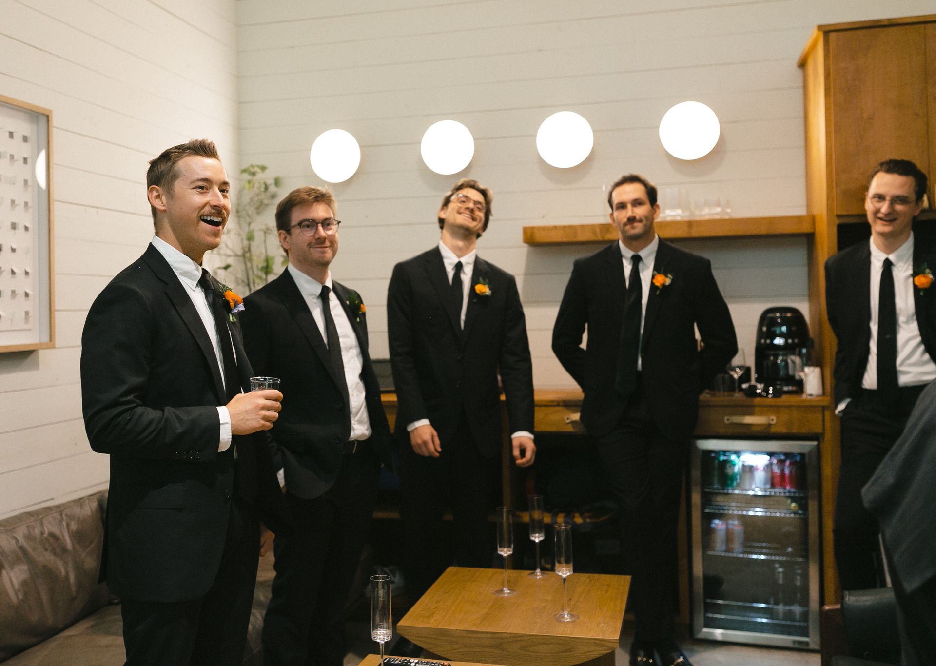 Men in black suits, celebratory moment. Some laugh, one tilts head back. Indoors, modern setting, grooms suite. 