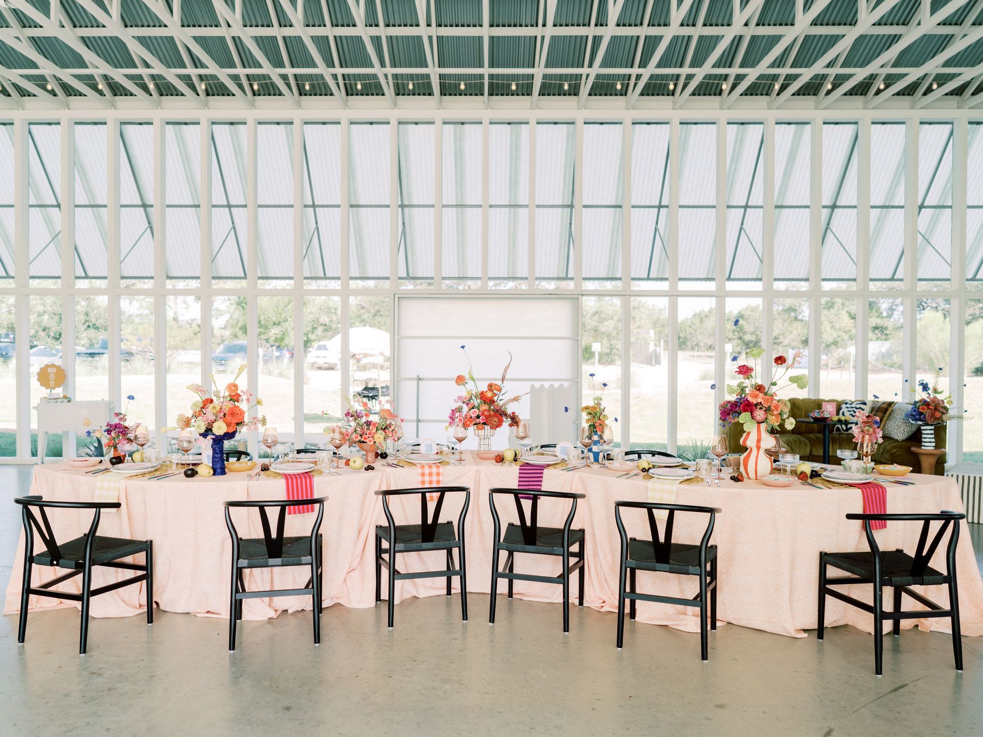 A table set with flowers and dinnerware is dressed with a blush tablecloth and black chairs around it in a large room with lots of windows.