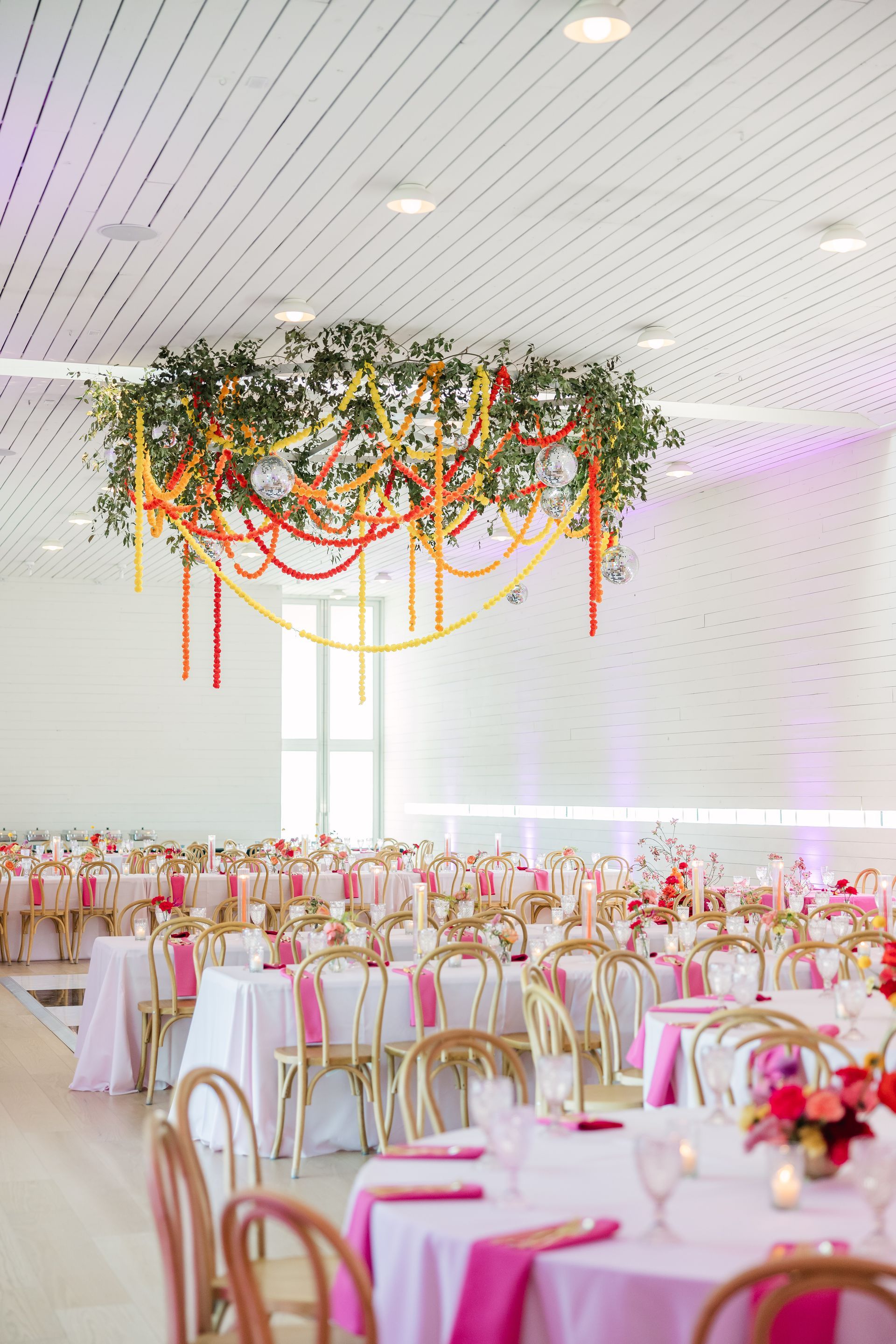 Wedding reception with pink and gold decor; a large floral installation hangs from the ceiling.