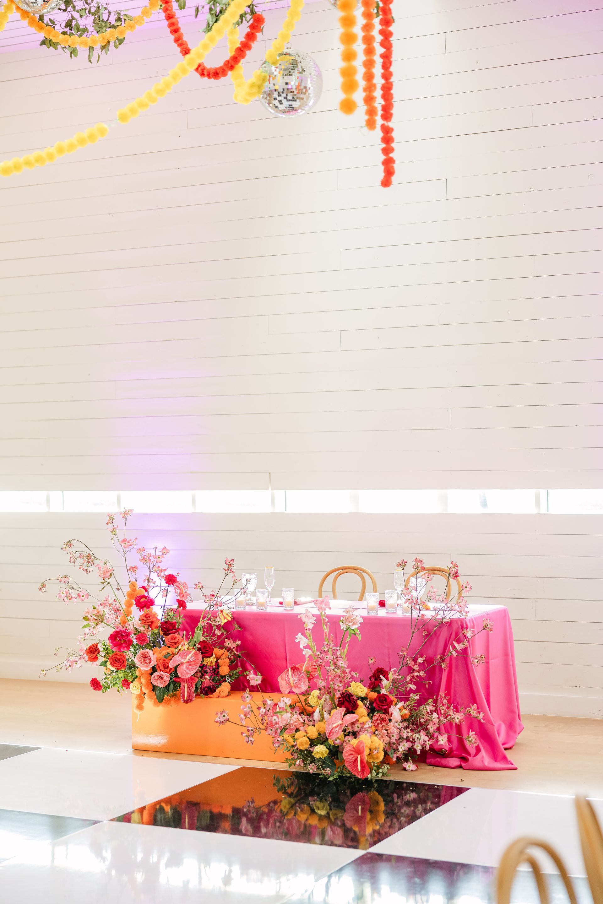 A wedding head table with floral arrangements, a bright pink tablecloth, and colorful decor hanging above.