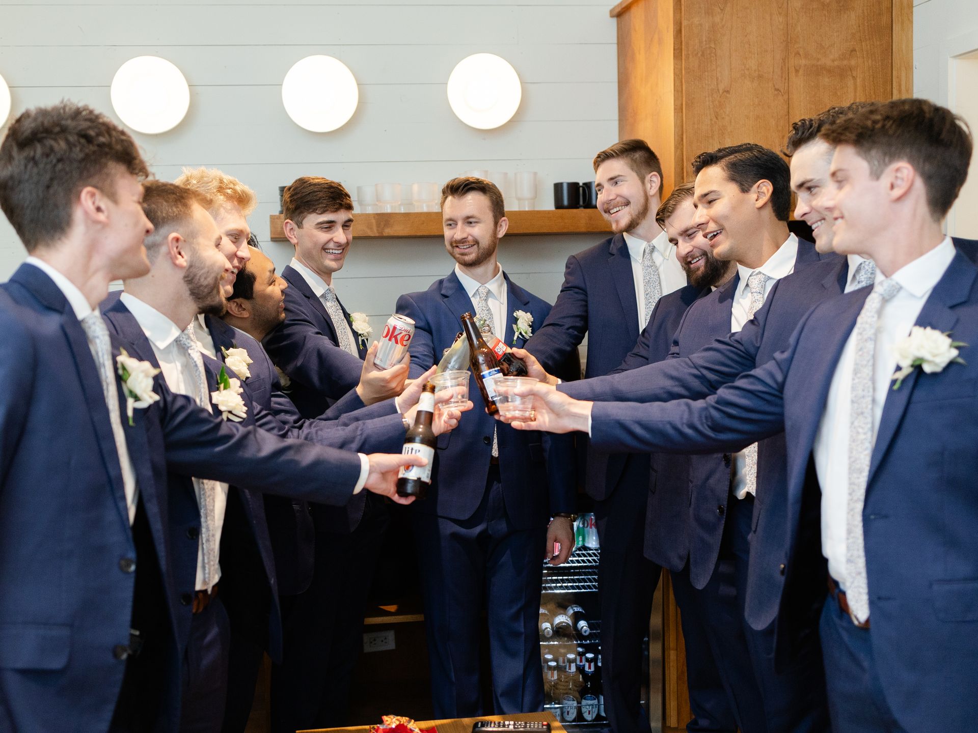 A group of men in navy suits are toasting with beer bottles and soda cans smiling at one another.