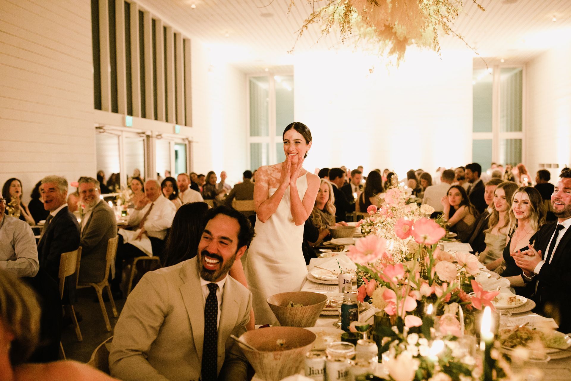 Bride applauds at wedding reception. Groom laughs beside floral centerpiece. Guests in background.