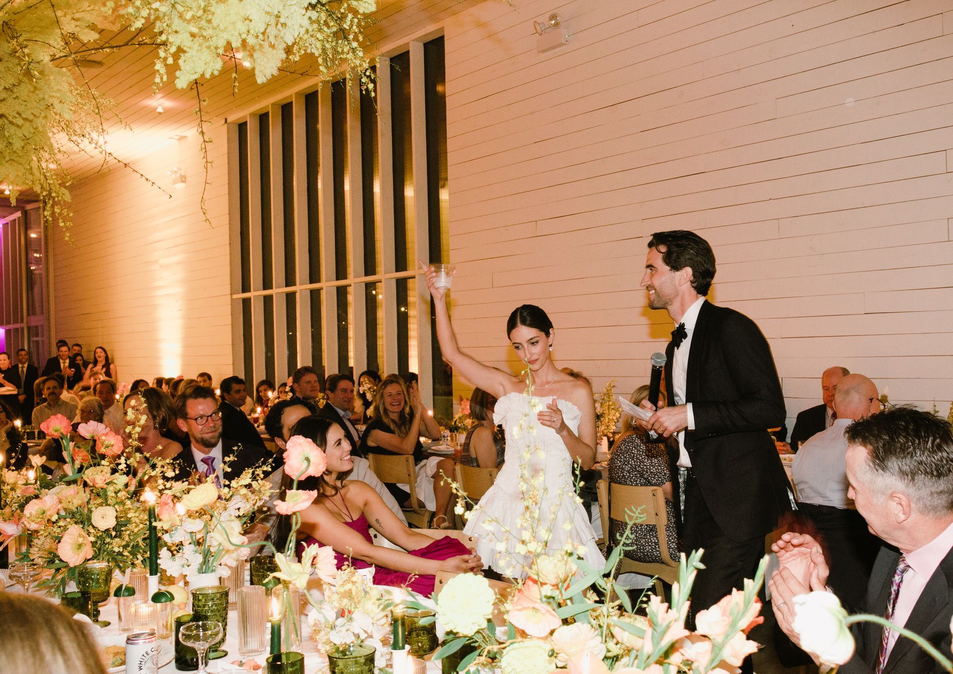 Wedding reception: Bride raises arm, groom speaks. Guests seated at tables with floral centerpieces.