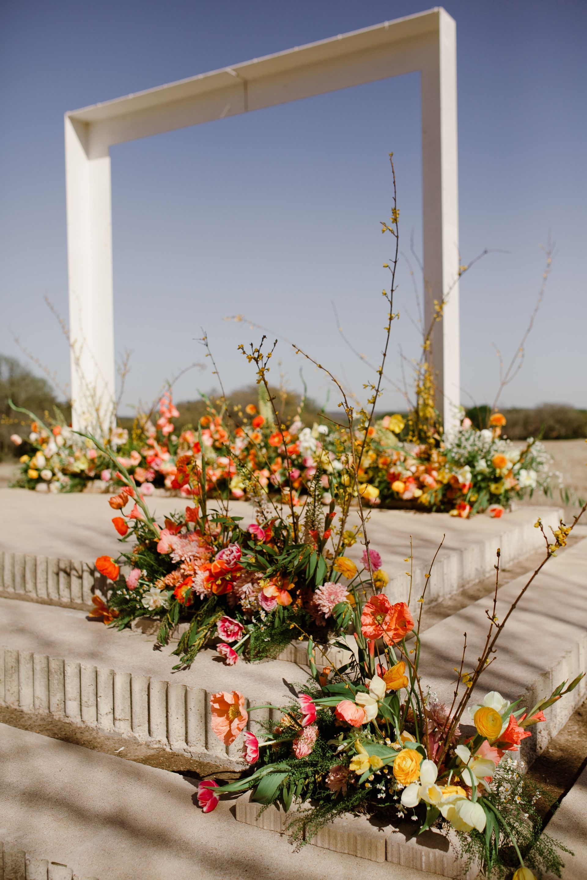 Floral arrangements cascade down steps towards a white rectangular frame, set against a clear sky at Prospect House in the Texas Hill Country. 