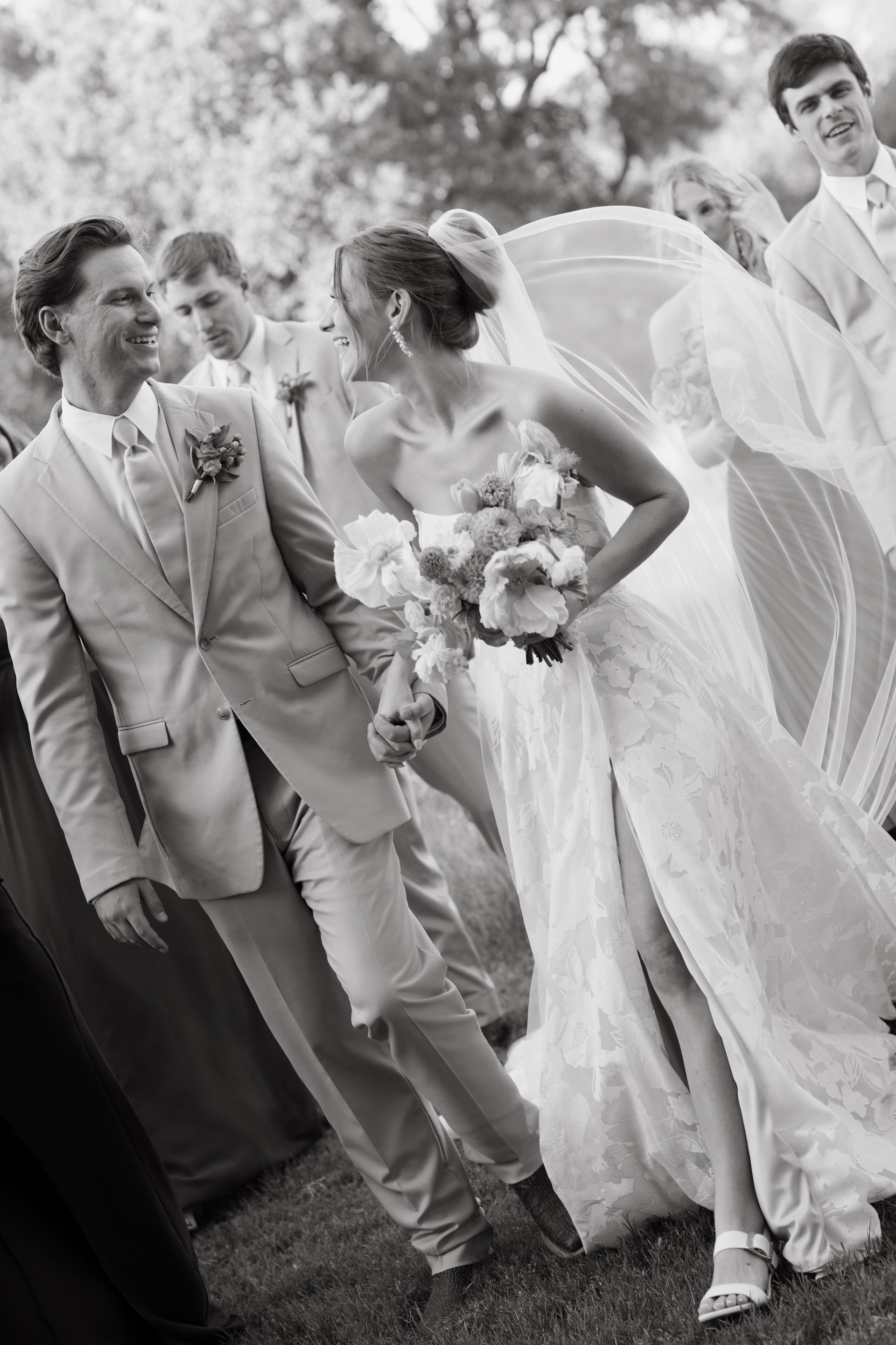 A black and white photo a bride and groom are laughing while walking and posing for a photo with their wedding party behind them.