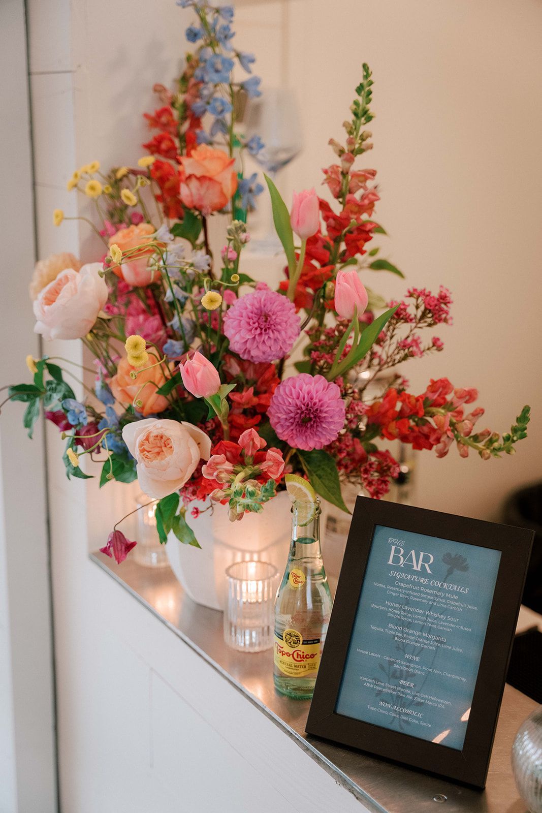 A bar top with a floral arrangement that is colorful with bright pinks, reds, yellows and greens. There is a bar menu to allow guest to review what is being served. 