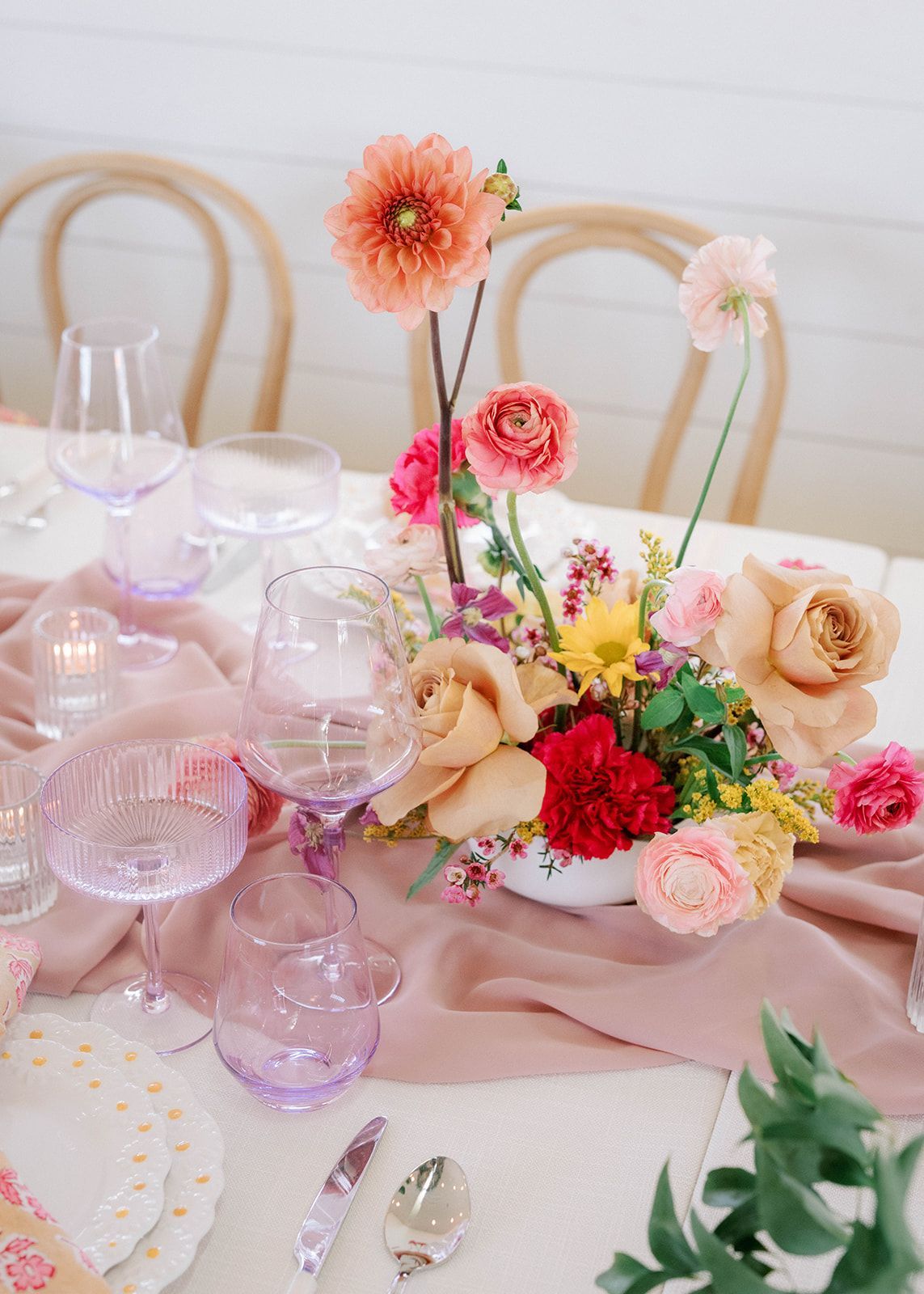 A table with a low vase filled with peach, yellow and ran flowers. The table is set with pale purple wine glasses and champange flutes.