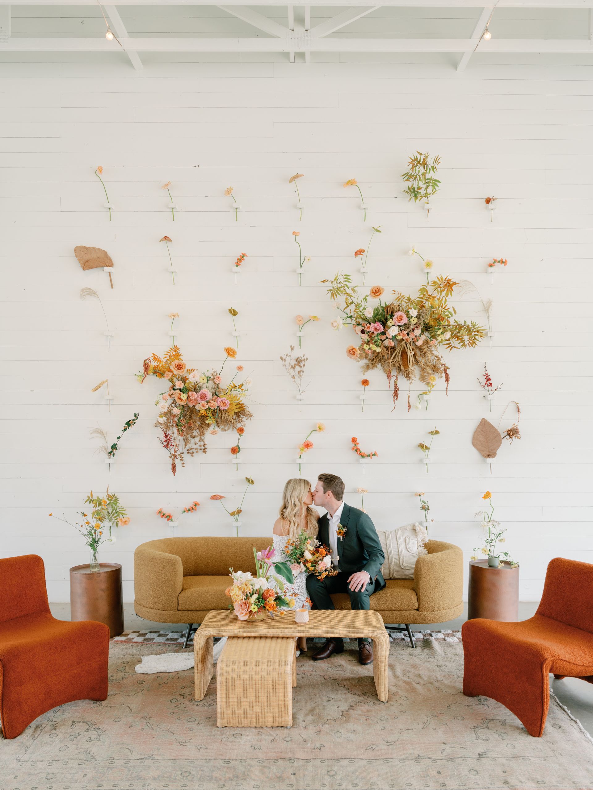 Couple kisses on mustard sofa, facing flower wall. Orange chairs flank the sofa.