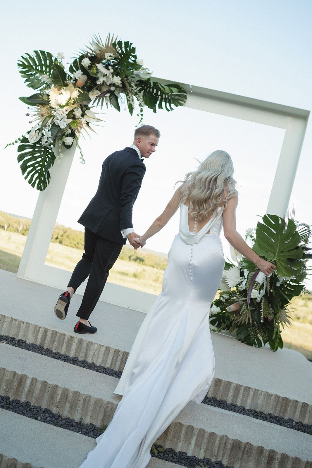 Bride and groom ascend stairs, holding hands. White dress, black suit, decorated arch, outdoor setting.