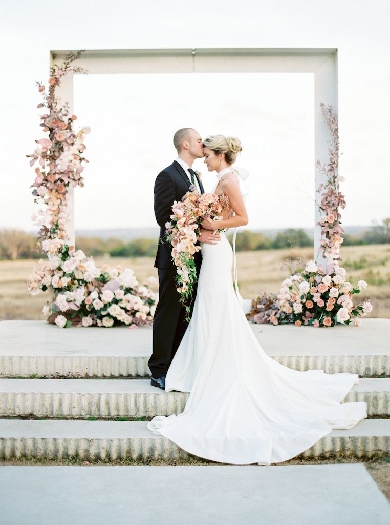 A bride and groom are kissing in front of a floral arch.