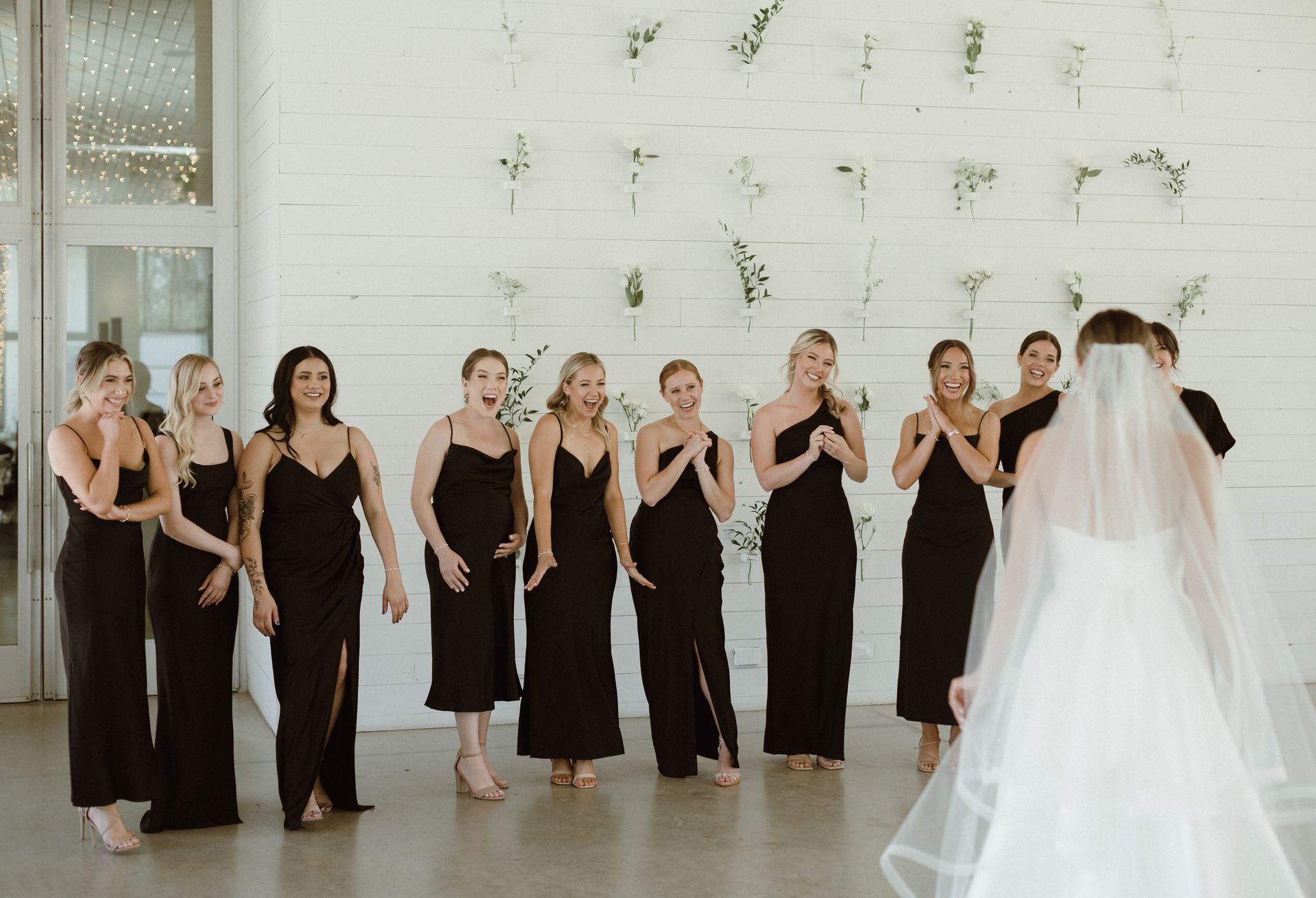 Bridesmaids in black dresses react to the bride in a white dress and veil; light-filled room.