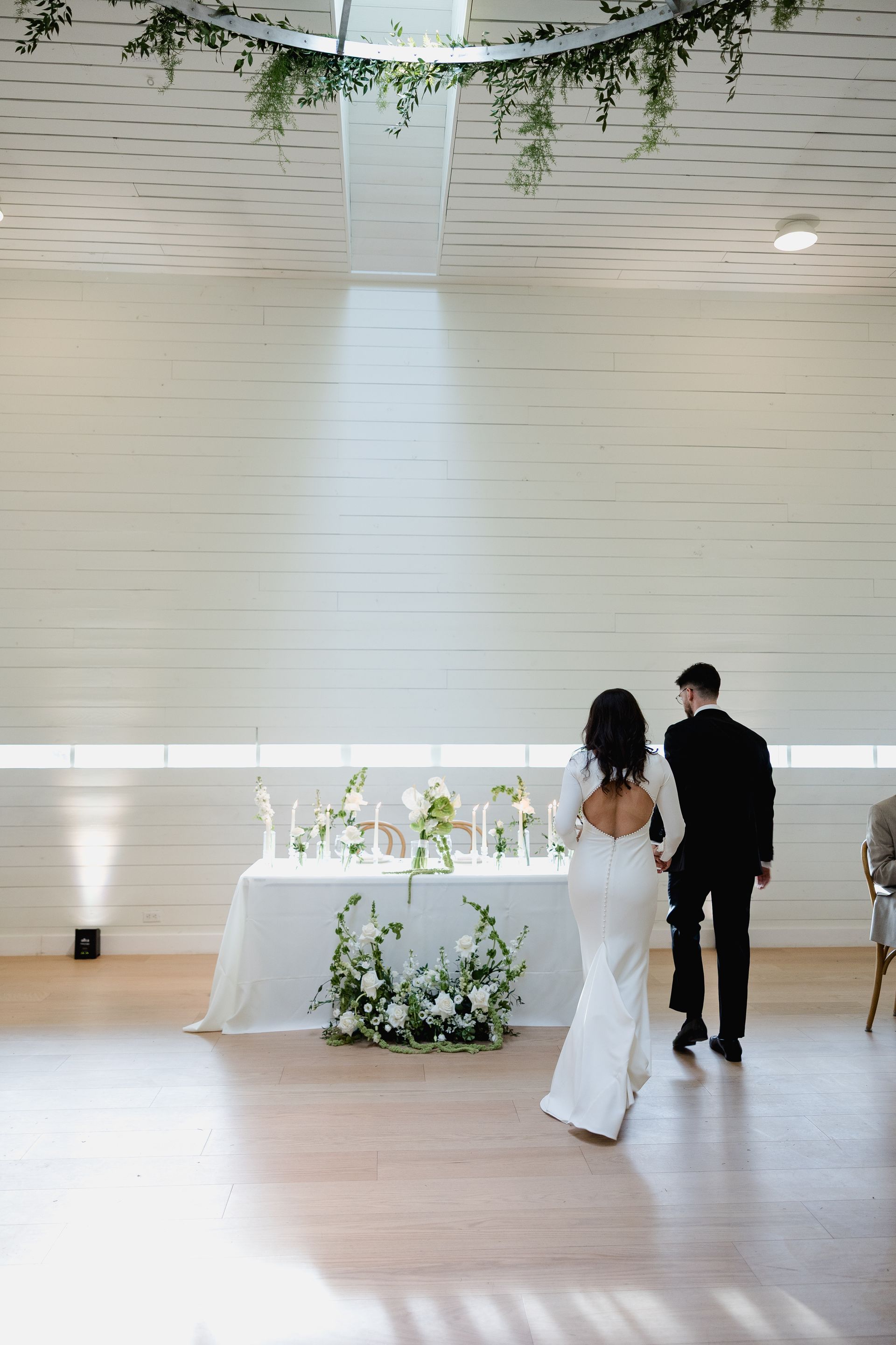 Bride and groom walk toward a table decorated with flowers, in a bright room.