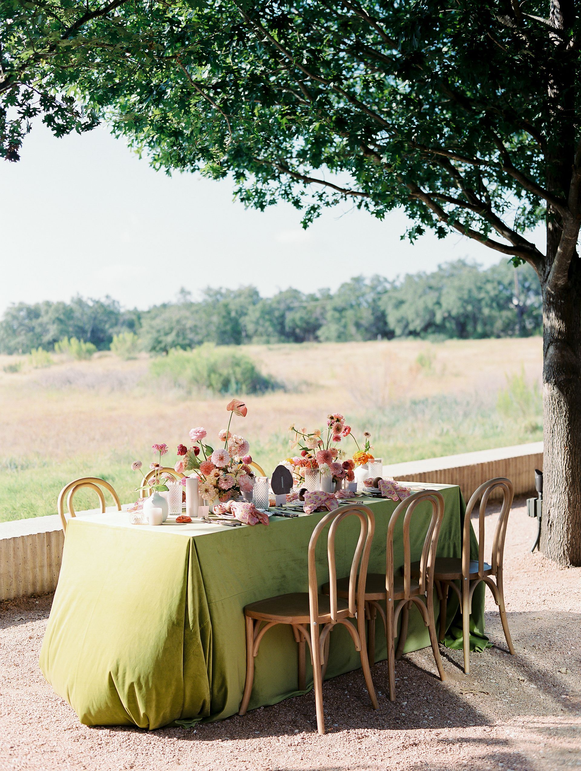 A group of people are gathering around round tables with gold linens on the side of a building surrounded by trees.