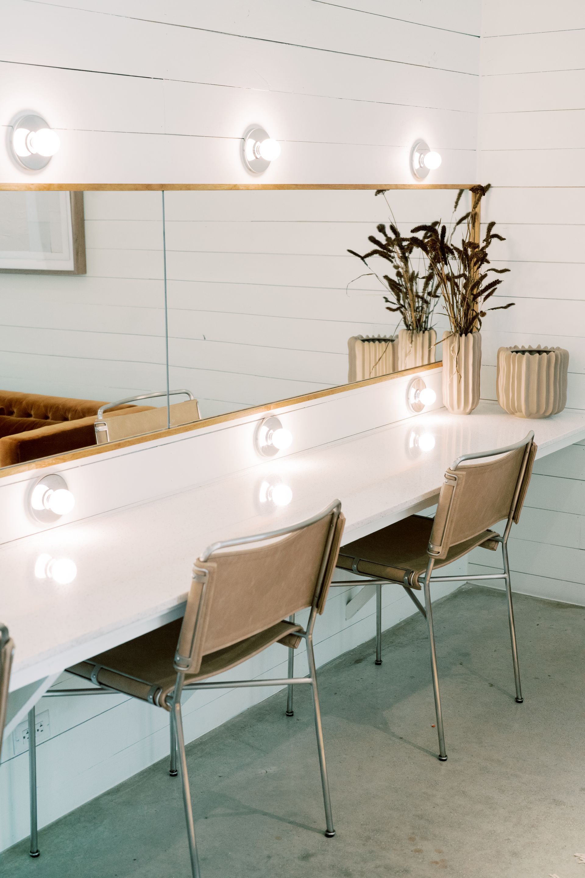 A dressing room with a long vanity with tan suede chairs and a large mirror with makeup lights. 