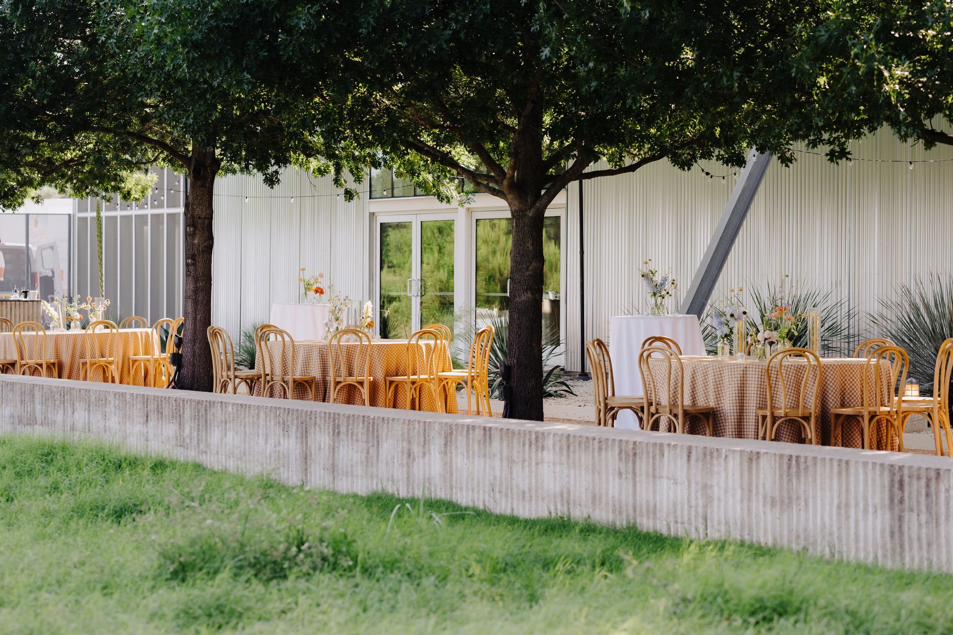Outdoor dining area with tables set for a gathering under trees. Yellow linens and wicker chairs.