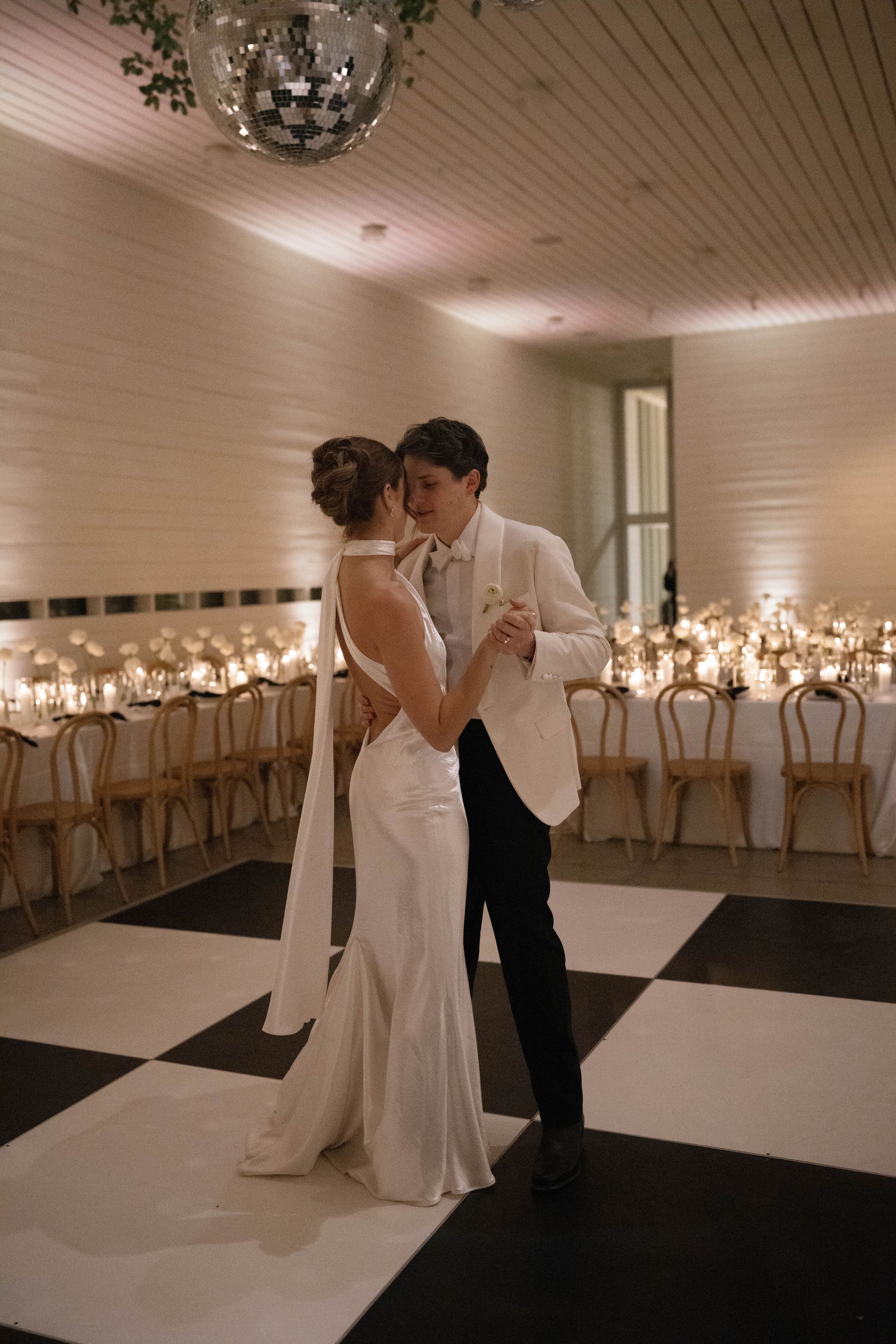 Couple dances on checkered floor, wedding reception. White gown and jacket, smiling, under a disco ball.