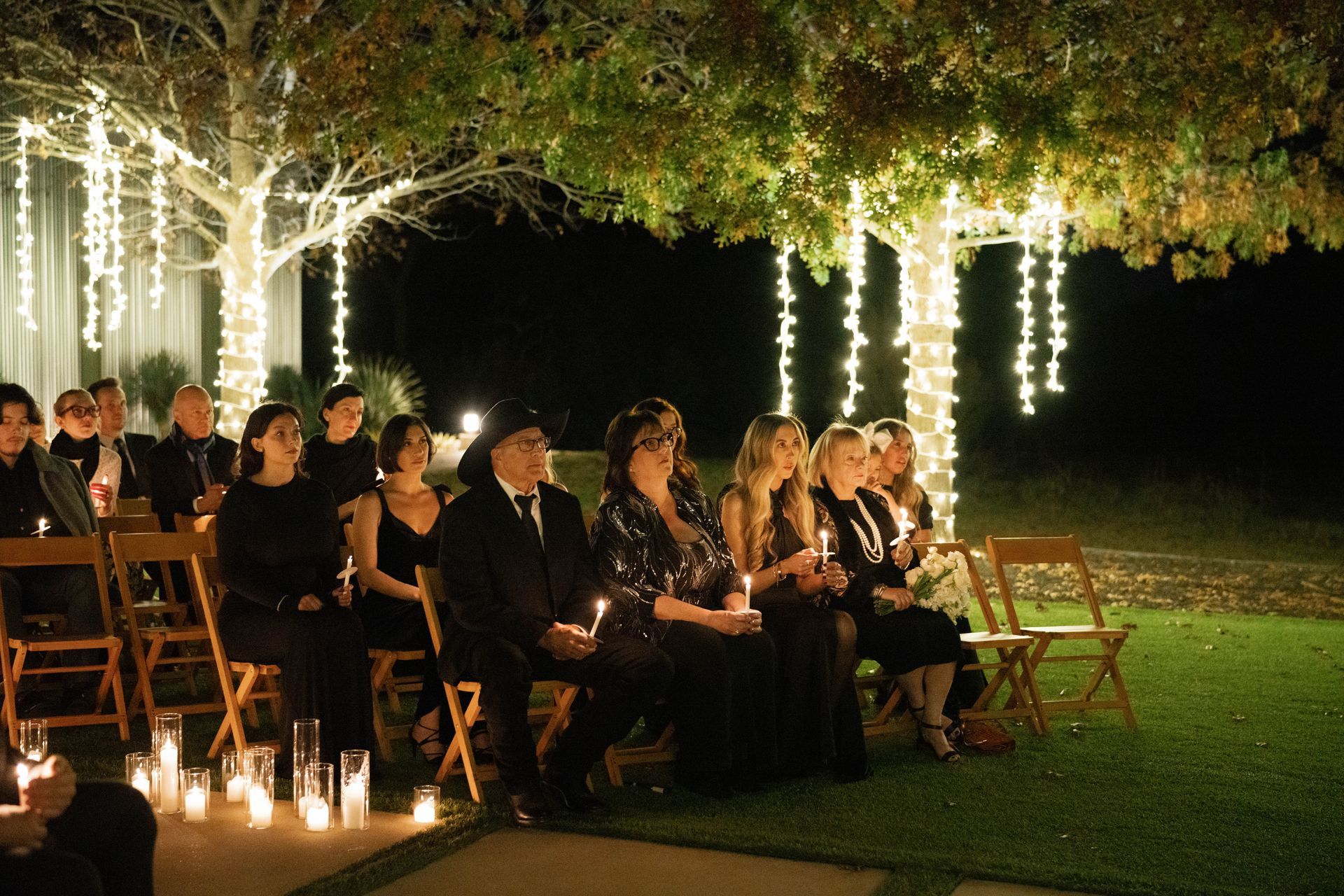 People seated outside at night, holding candles, under trees with string lights.