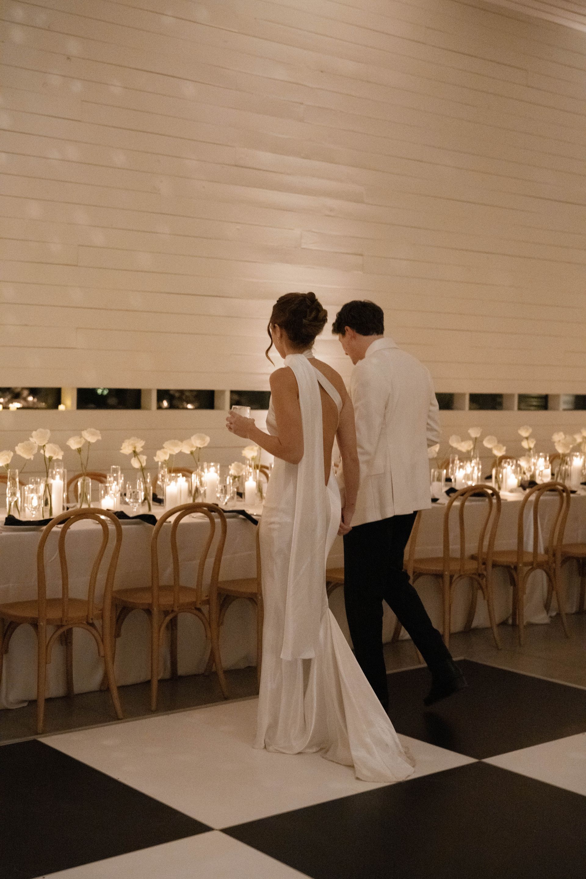 Couple walking towards a table with candles and flowers, in a white-walled room with a checkered floor.