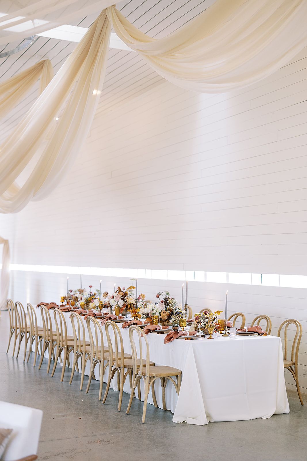 A long table in a white room with wood arched chairs and flowers scattered in the middle with candles throughout. 