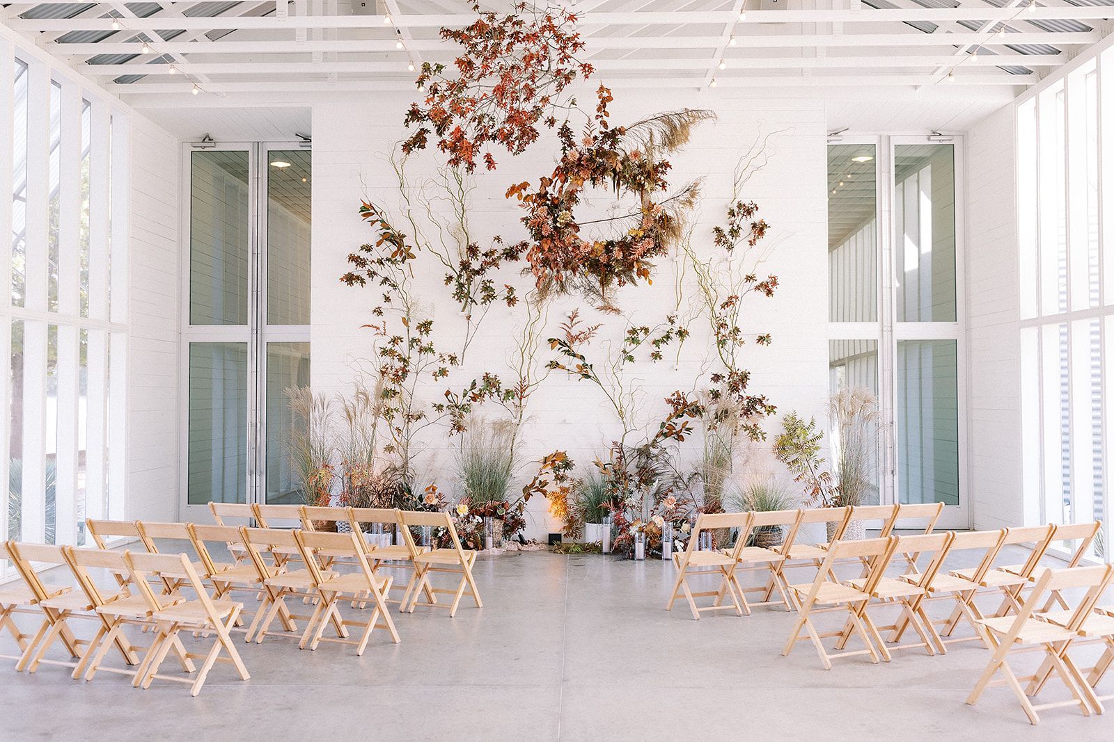 A few rows of wooden folding chairs are set up in front of a white wall with fall leaves and flowers on it and an aisle between the two sets of rows. 