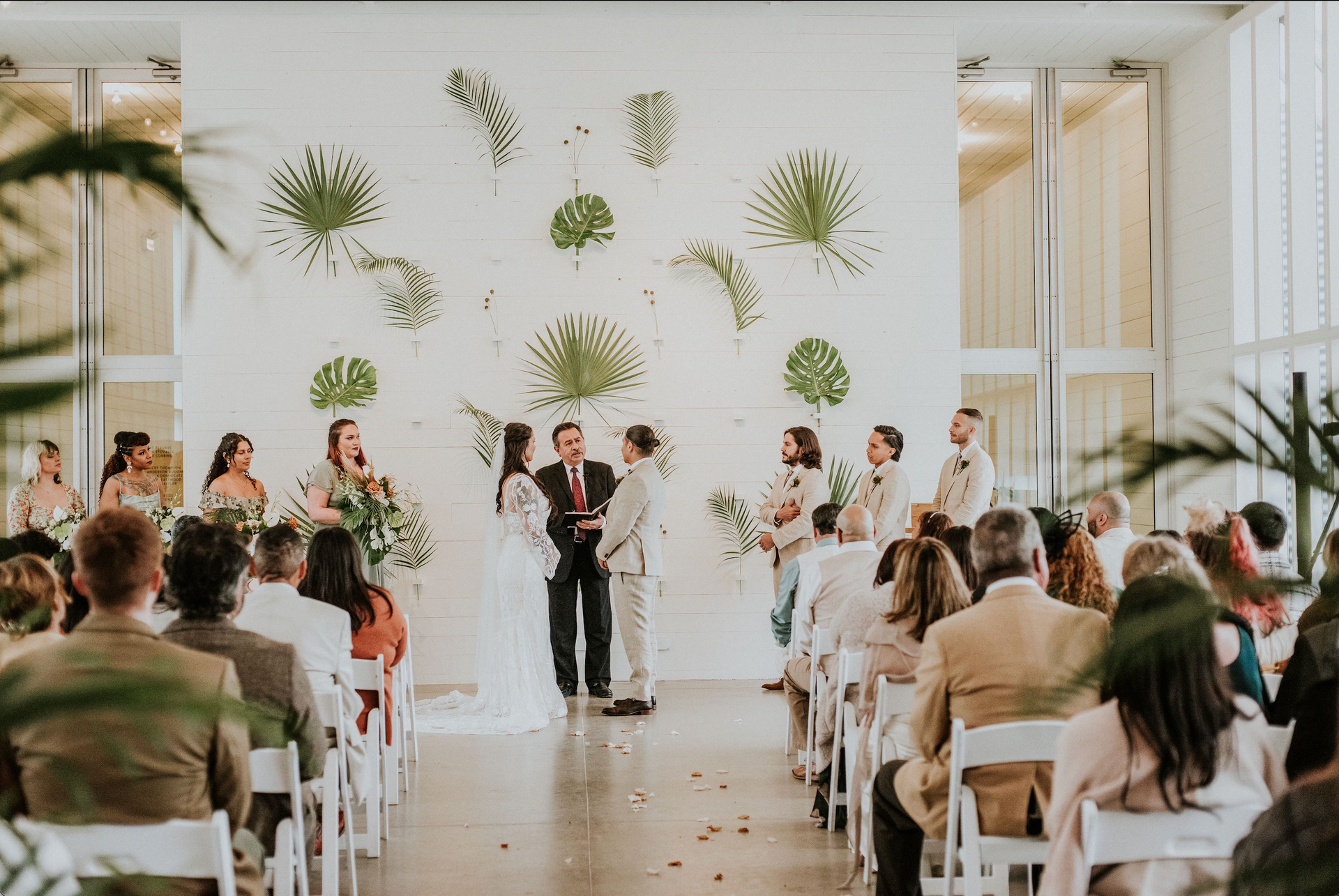 Wedding ceremony with couple, officiant, and guests in a white room with palm leaves.