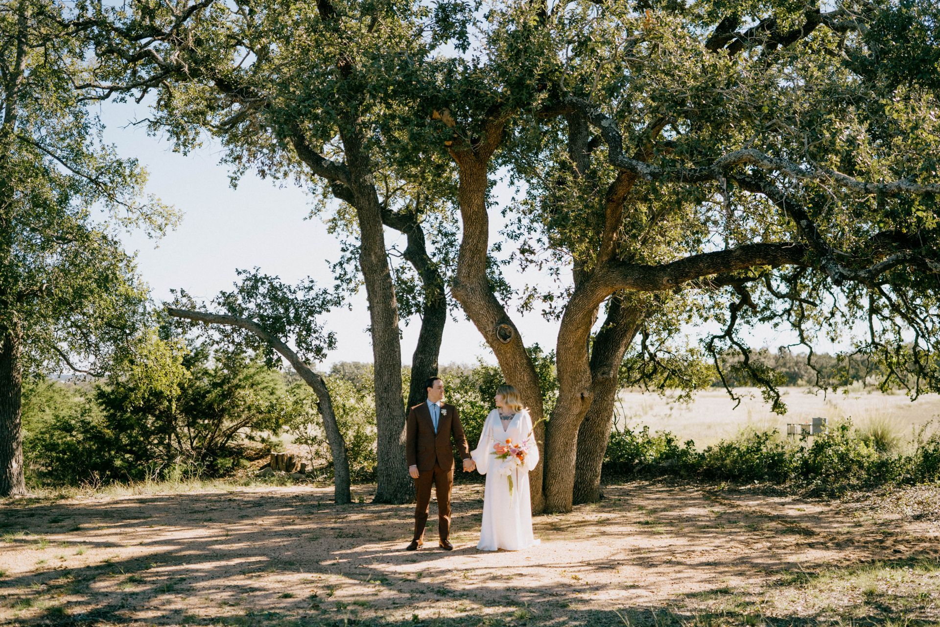 A bride in a white long sleeve dress and groom in a brown suit are standing under a tree in the dirt.