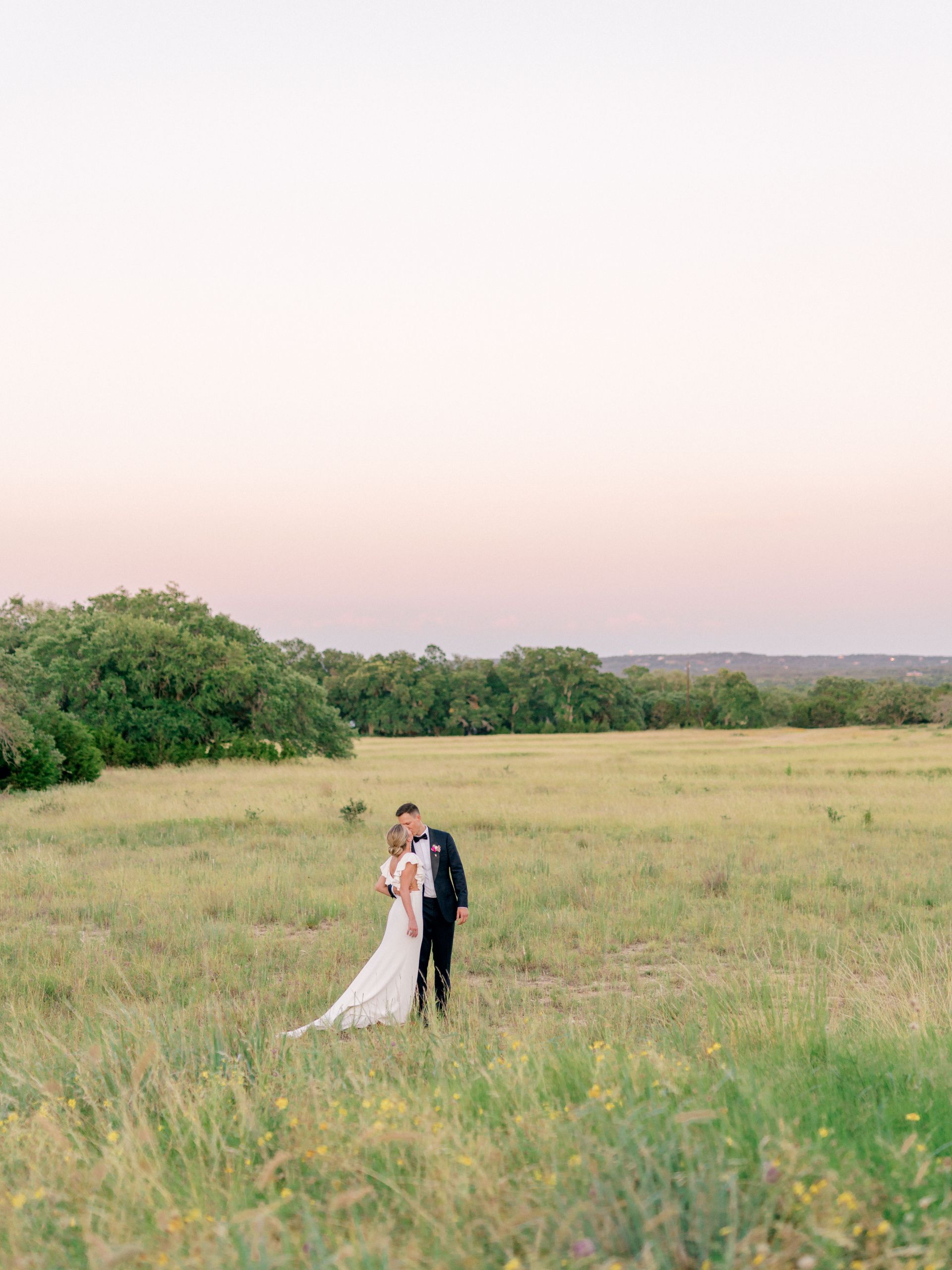 A bride and groom are standing in a field with a pale pink toned sunset in the sky. 