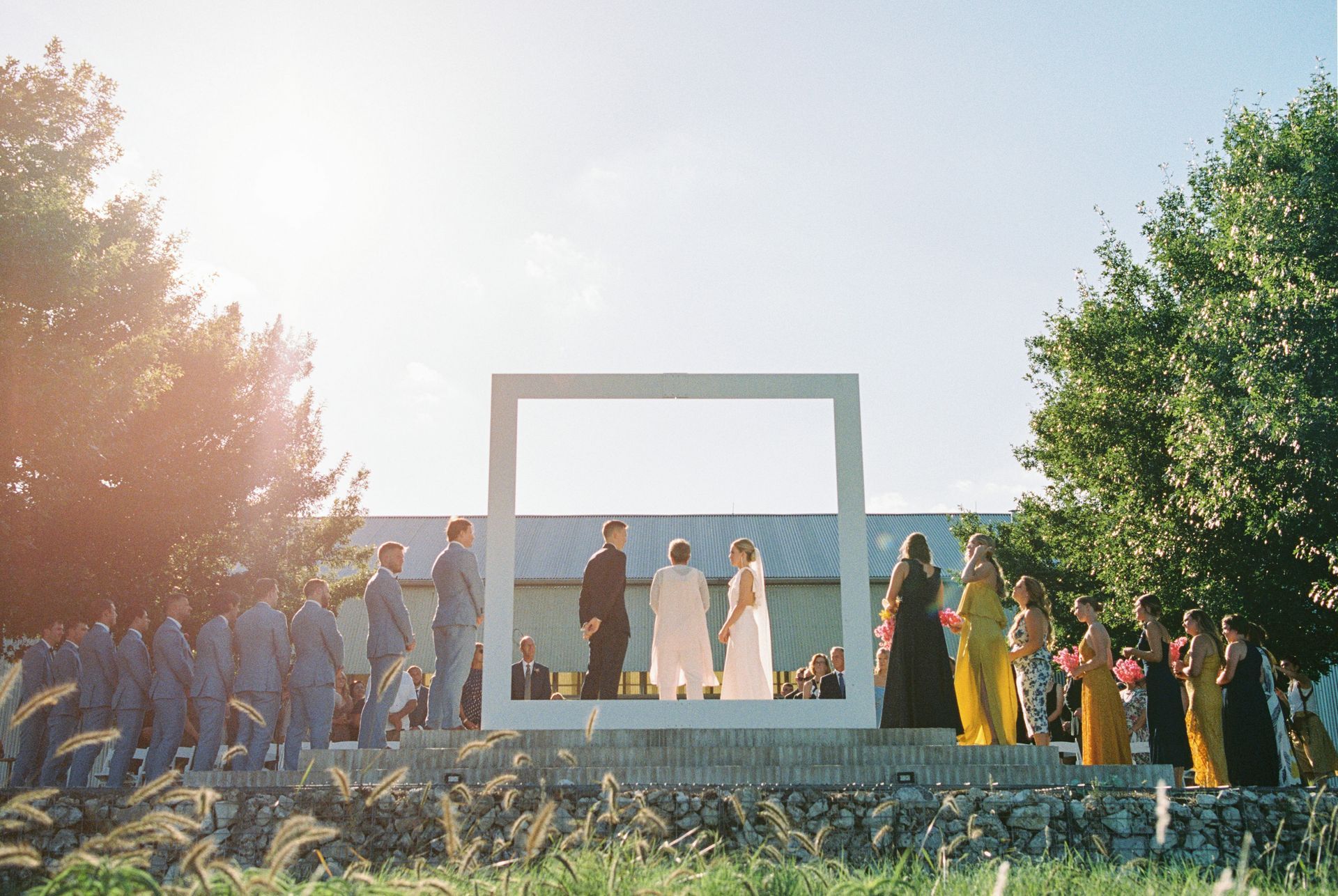Wedding ceremony: couple in front of a large white frame, surrounded by wedding party in a sunny outdoor setting.