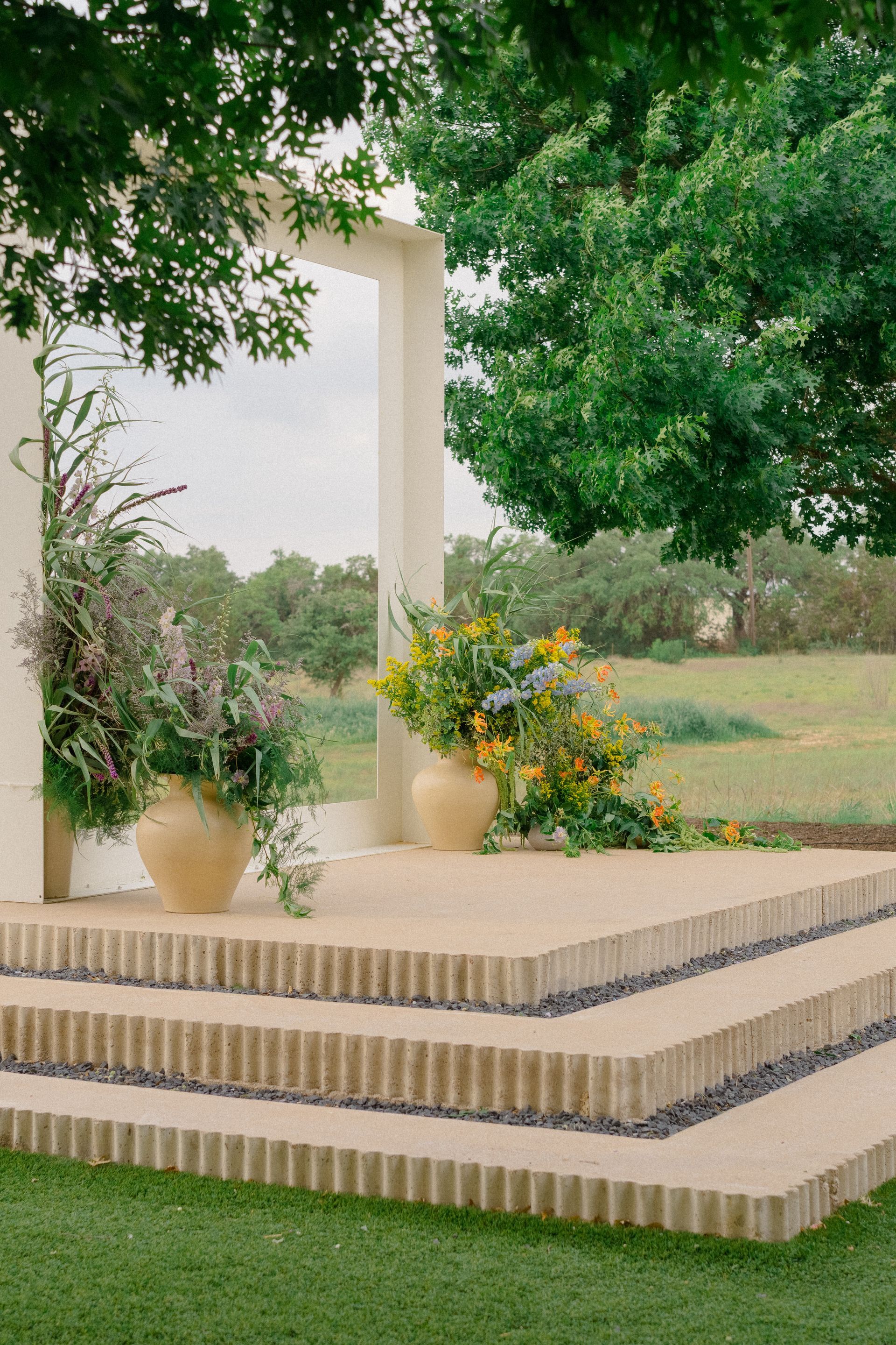 Stone steps lead to an outdoor stage with floral arrangements. Green grass and trees surround.