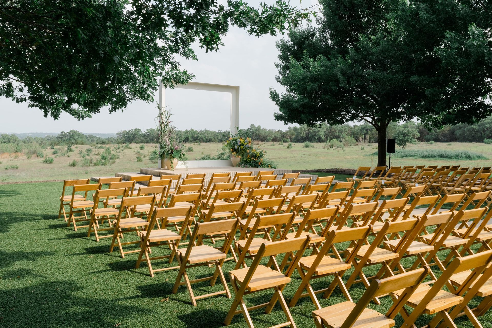 Rows of wooden chairs face a simple altar beneath trees, in a grassy outdoor ceremony.
