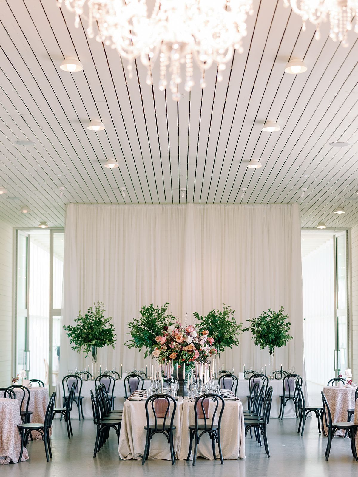 Elegant wedding reception with floral centerpieces and draped backdrop. Black chairs surround tables.