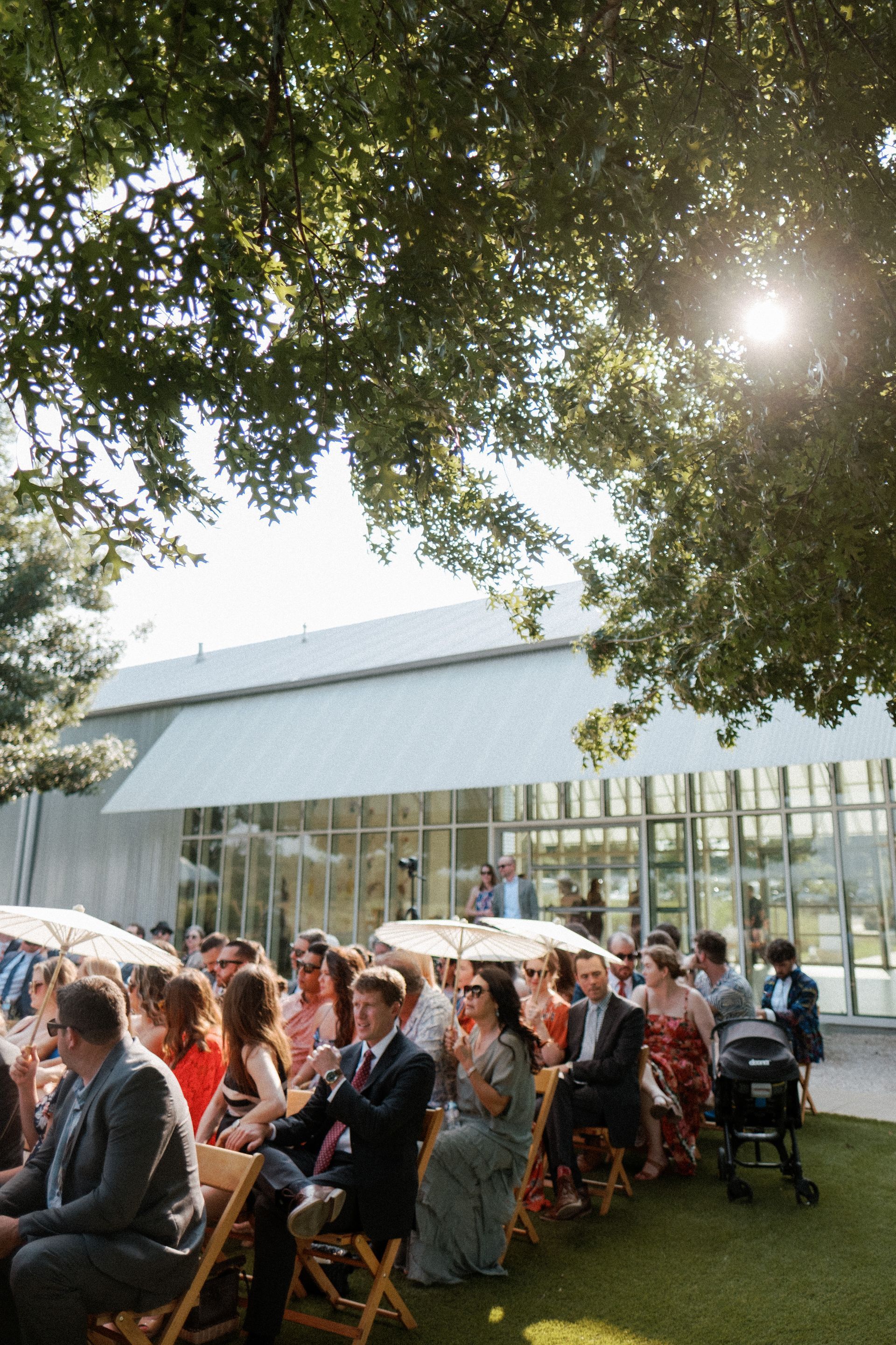 A bride and groom are walking down a sidewalk holding hands while the groom is holding her lush green and white bouquet.