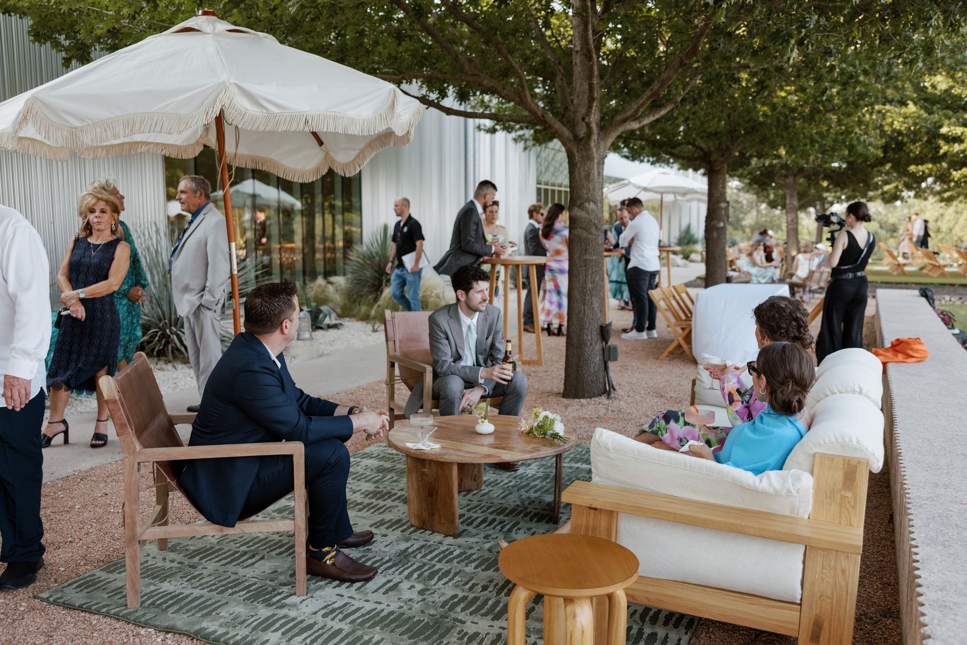 Guests mingle outdoors at a social event. People dressed in formal attire converse near furniture under trees.
